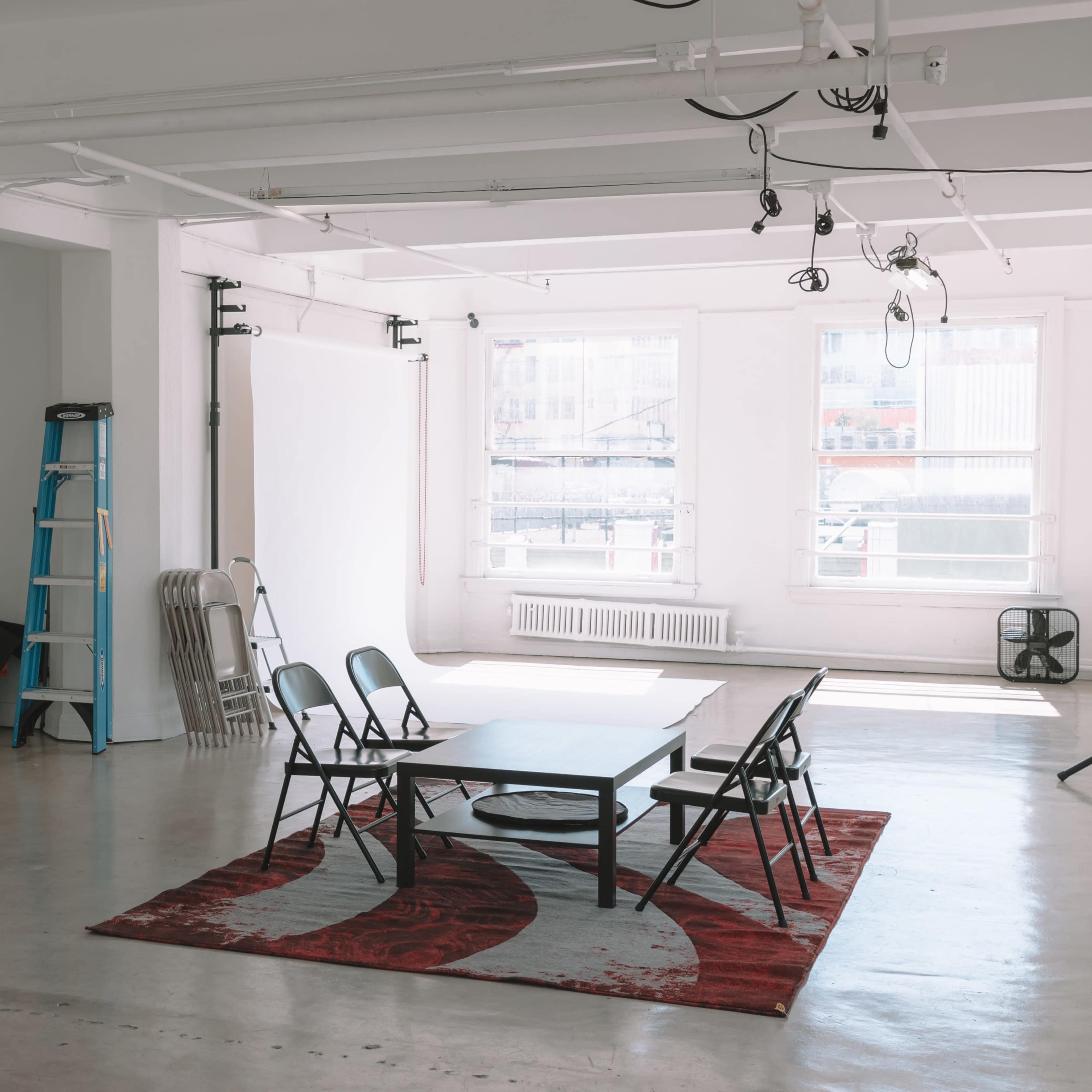 An empty studio space features a table surrounded by black chairs, a large carpet, and various equipment along the walls.