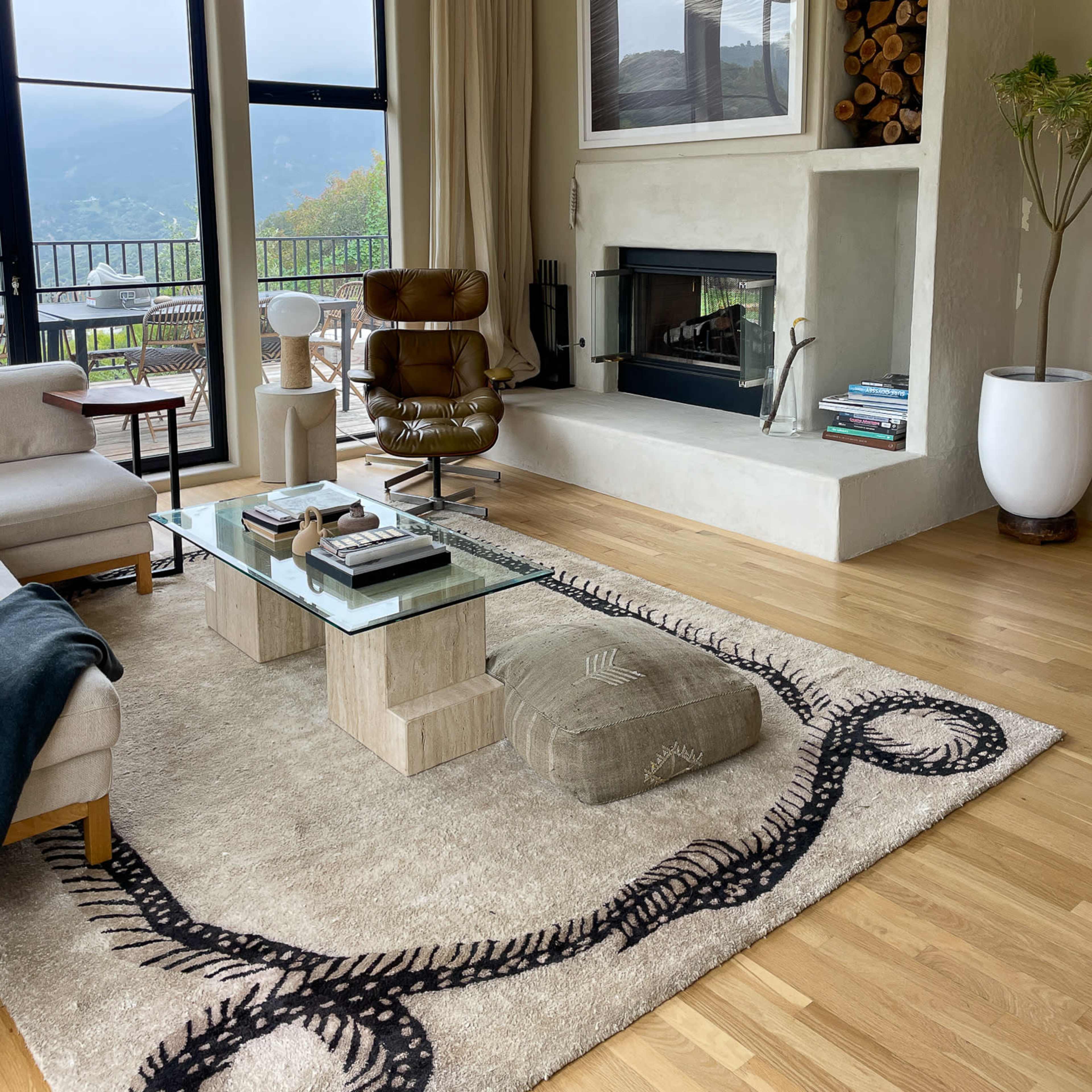 The living room features a fireplace with stacked logs, a glass coffee table, a comfortable chair, and large windows overlooking a mountainous view.