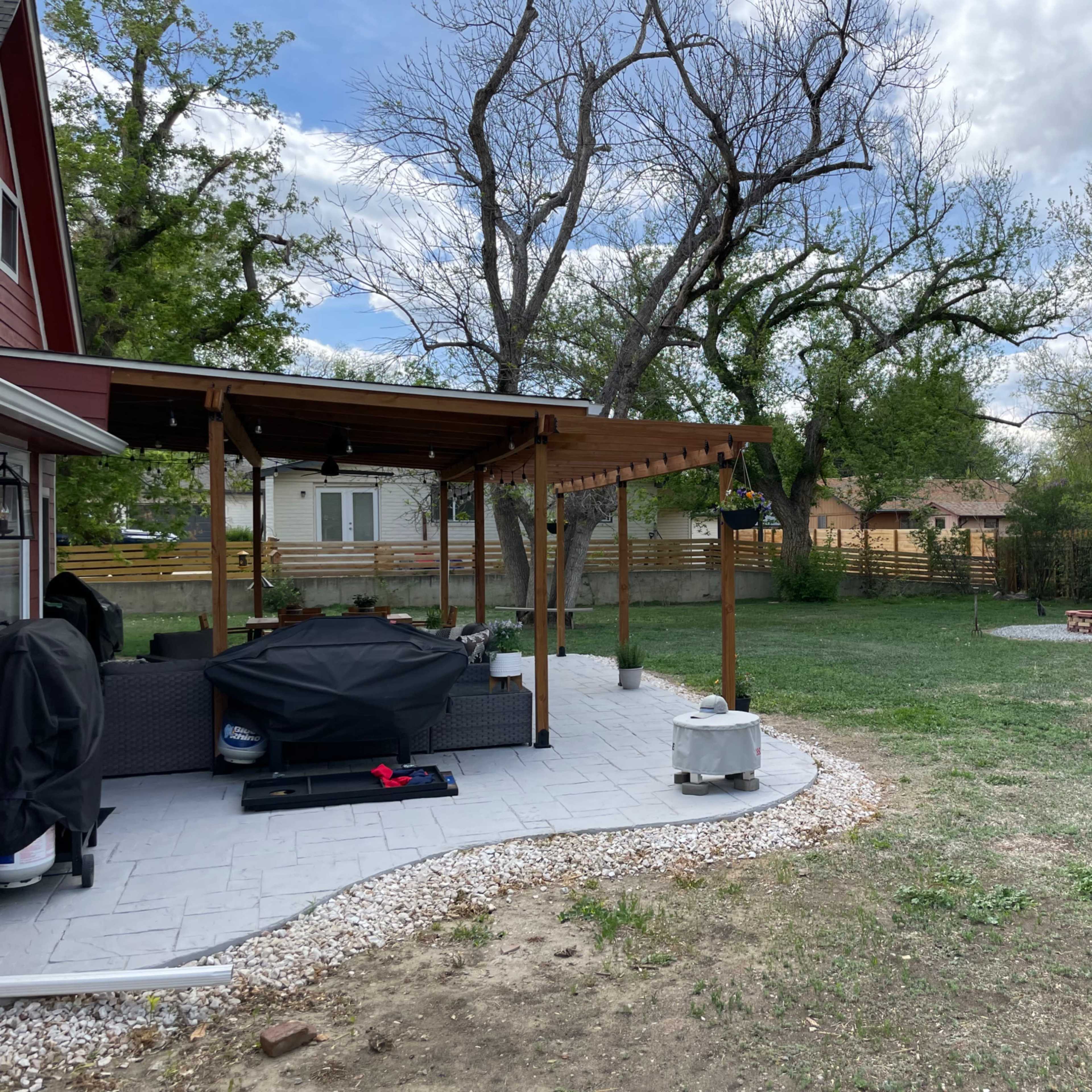 A backyard with a paved patio area covered by a wooden pergola, featuring outdoor furniture and a grill, while an open lawn and trees are visible in the background.