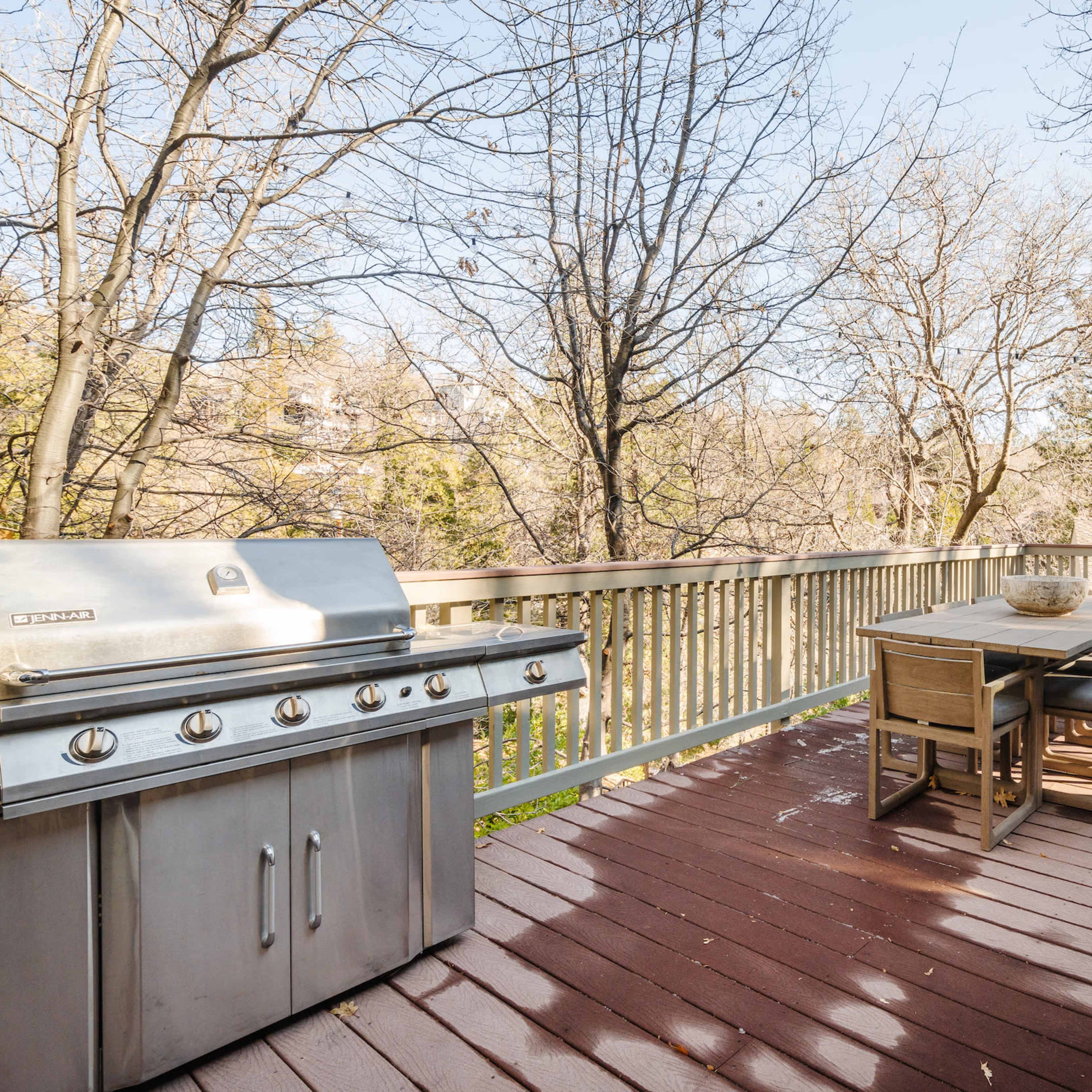A wooden deck features a stainless steel grill and a dining table with chairs, surrounded by trees in a residential area.