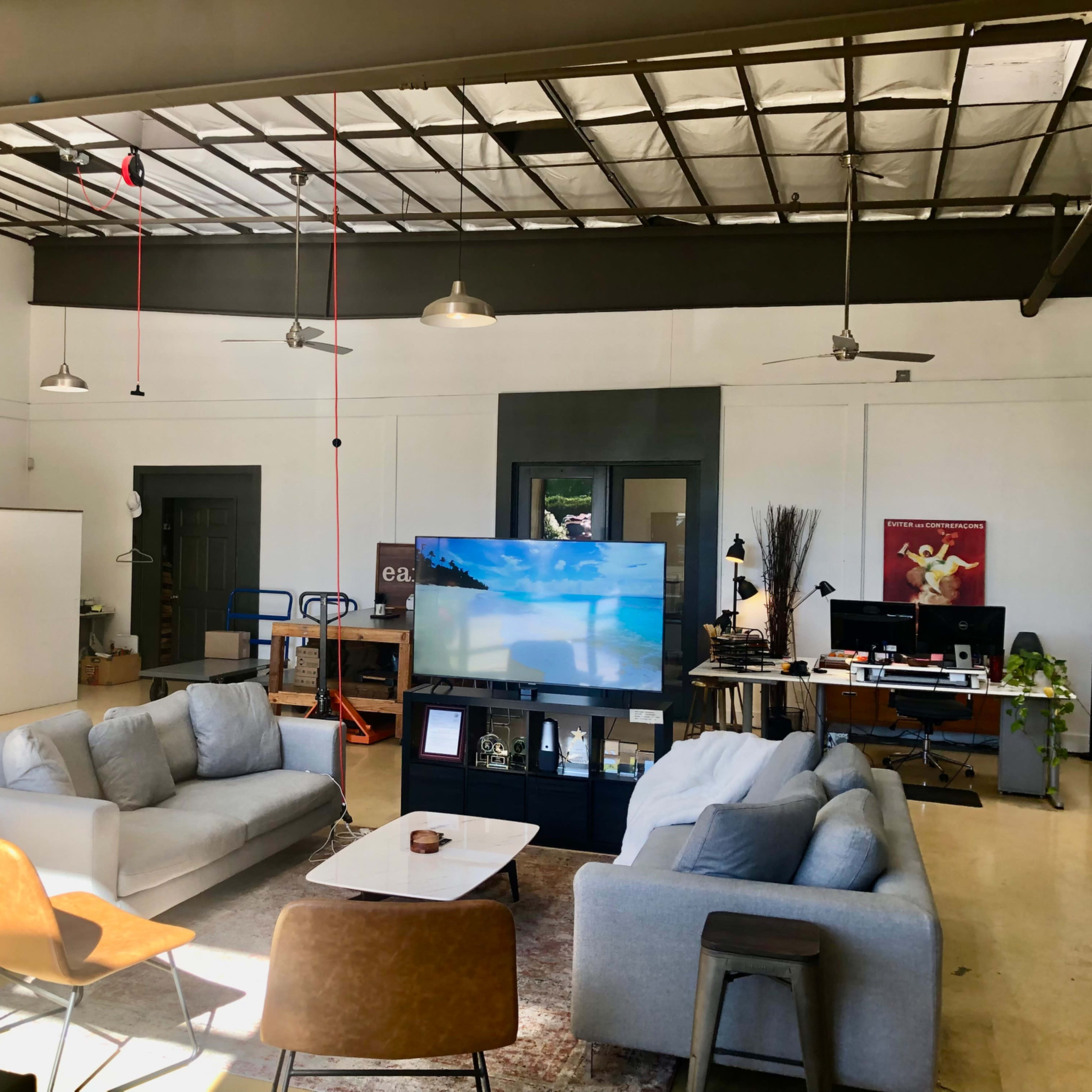 A spacious office interior with modern furniture, including two sofas and a television, surrounded by workstations and desk chairs.