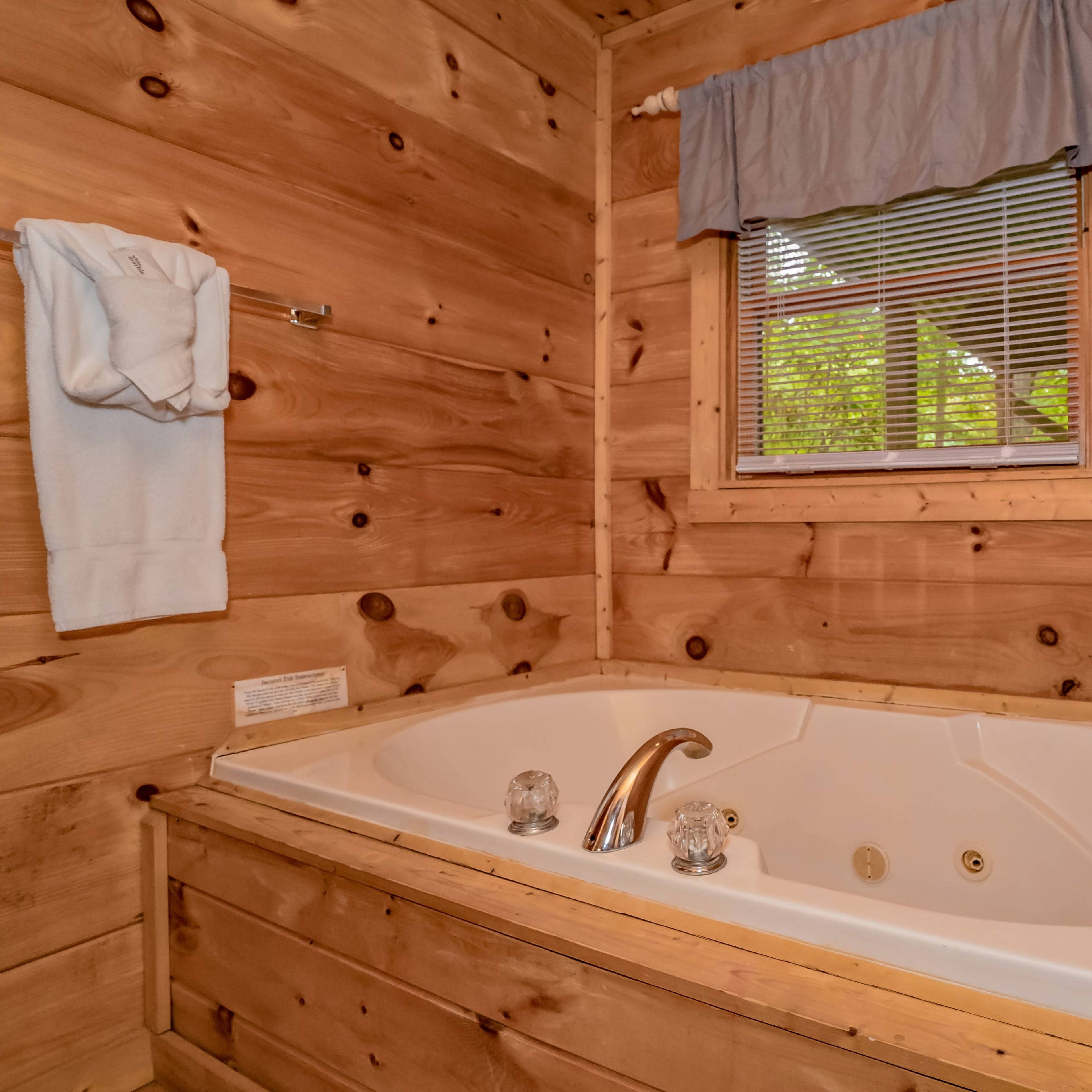 A wooden bathroom featuring a large white jacuzzi tub beneath a window with a light blue curtain.