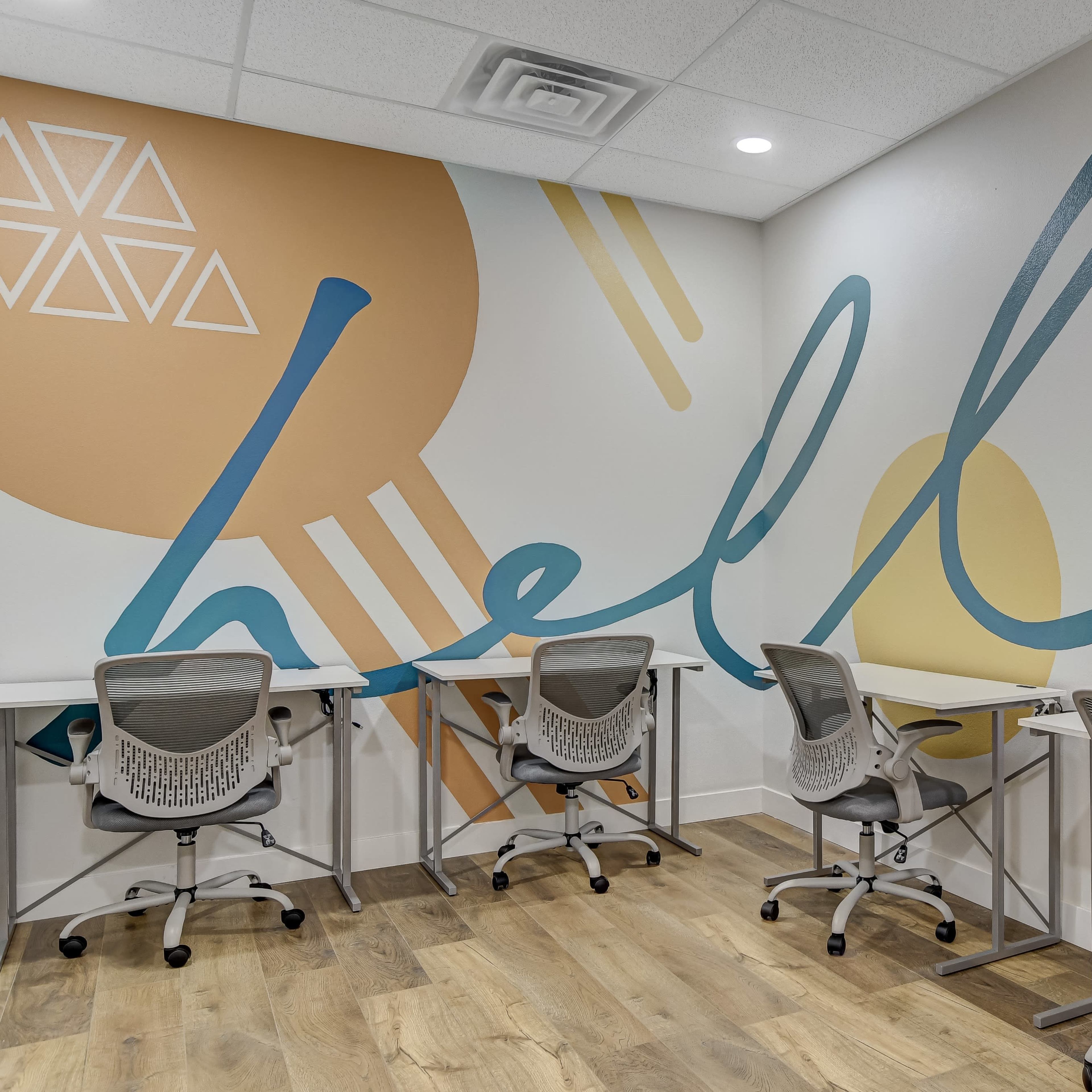 A modern workspace with four chairs and desks arranged against a wall featuring a colorful mural that includes the word "hello."