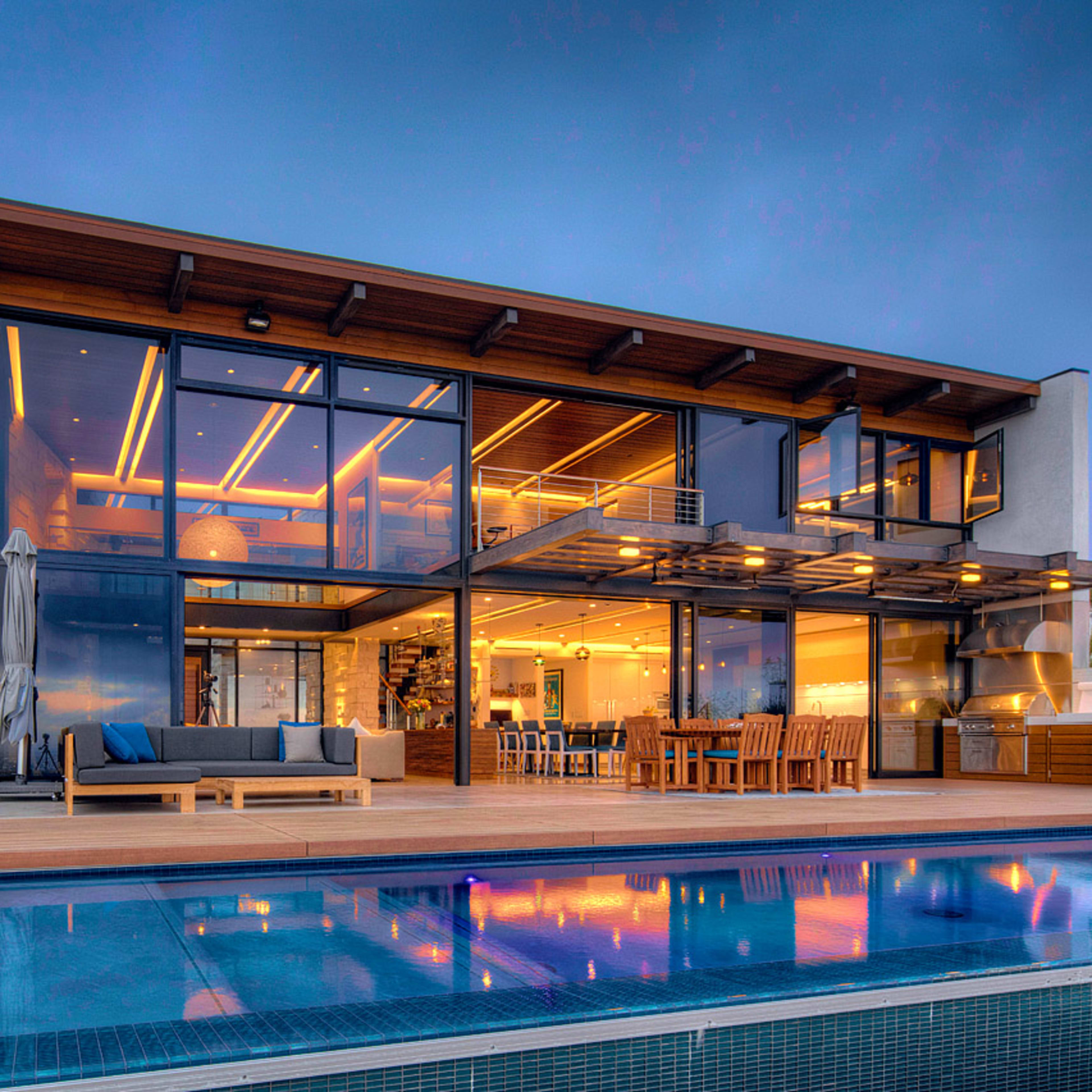 A modern two-story house features large glass windows and a patio by a swimming pool.