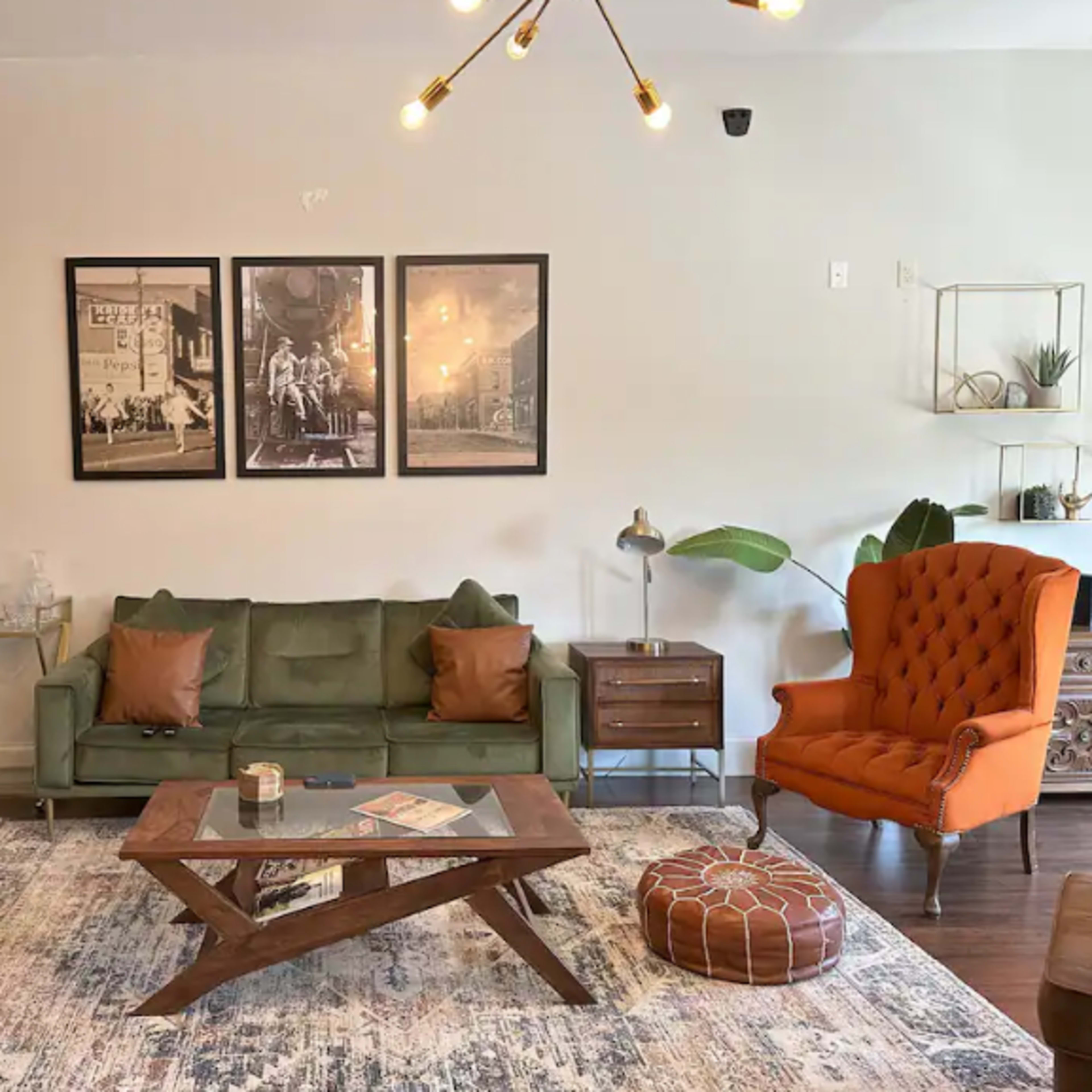 A living room is depicted with a green sofa, an orange armchair, a wooden coffee table, and framed artwork on the wall.