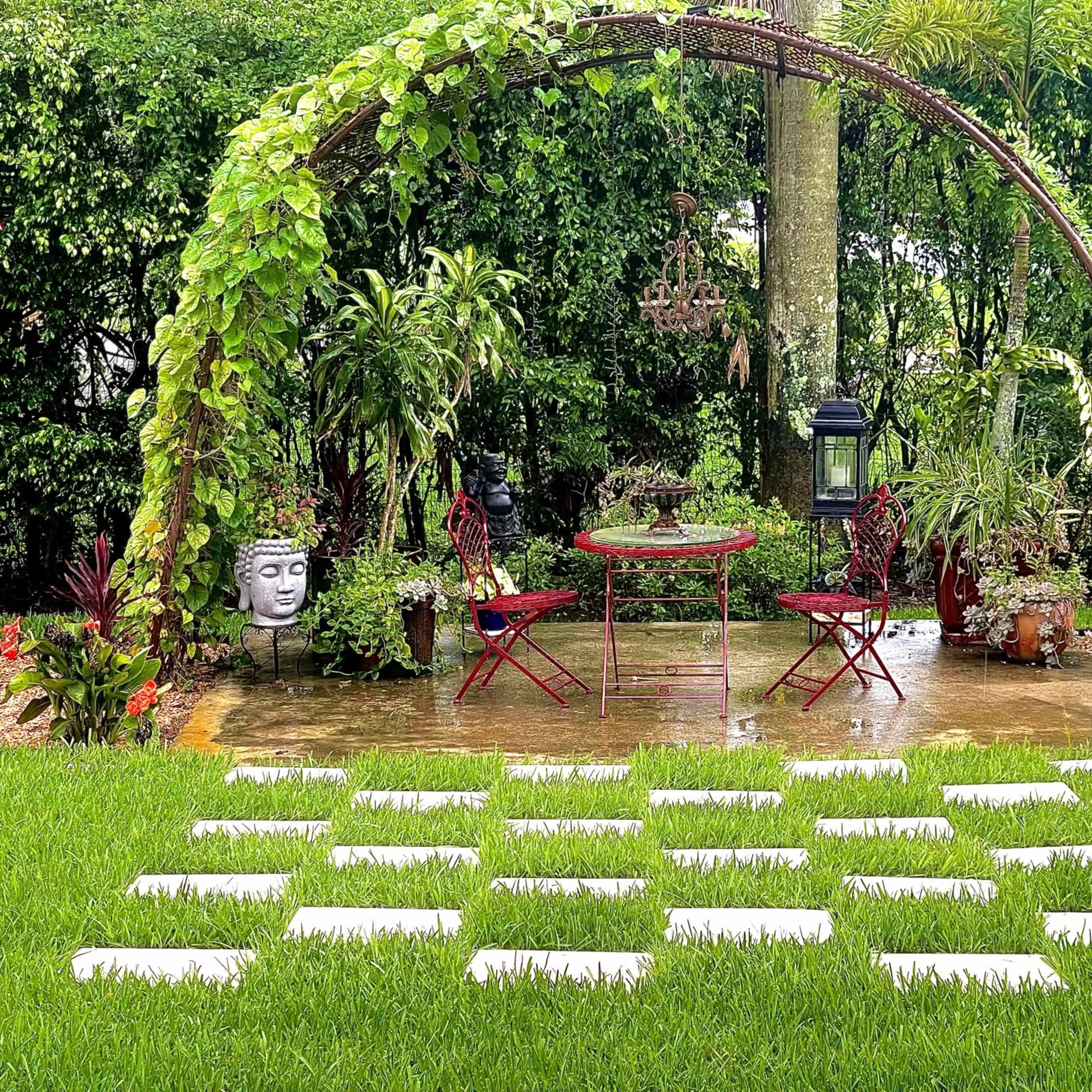 A quaint outdoor seating area with a round table and two red chairs is surrounded by lush greenery and decorative plants under a metal arch.