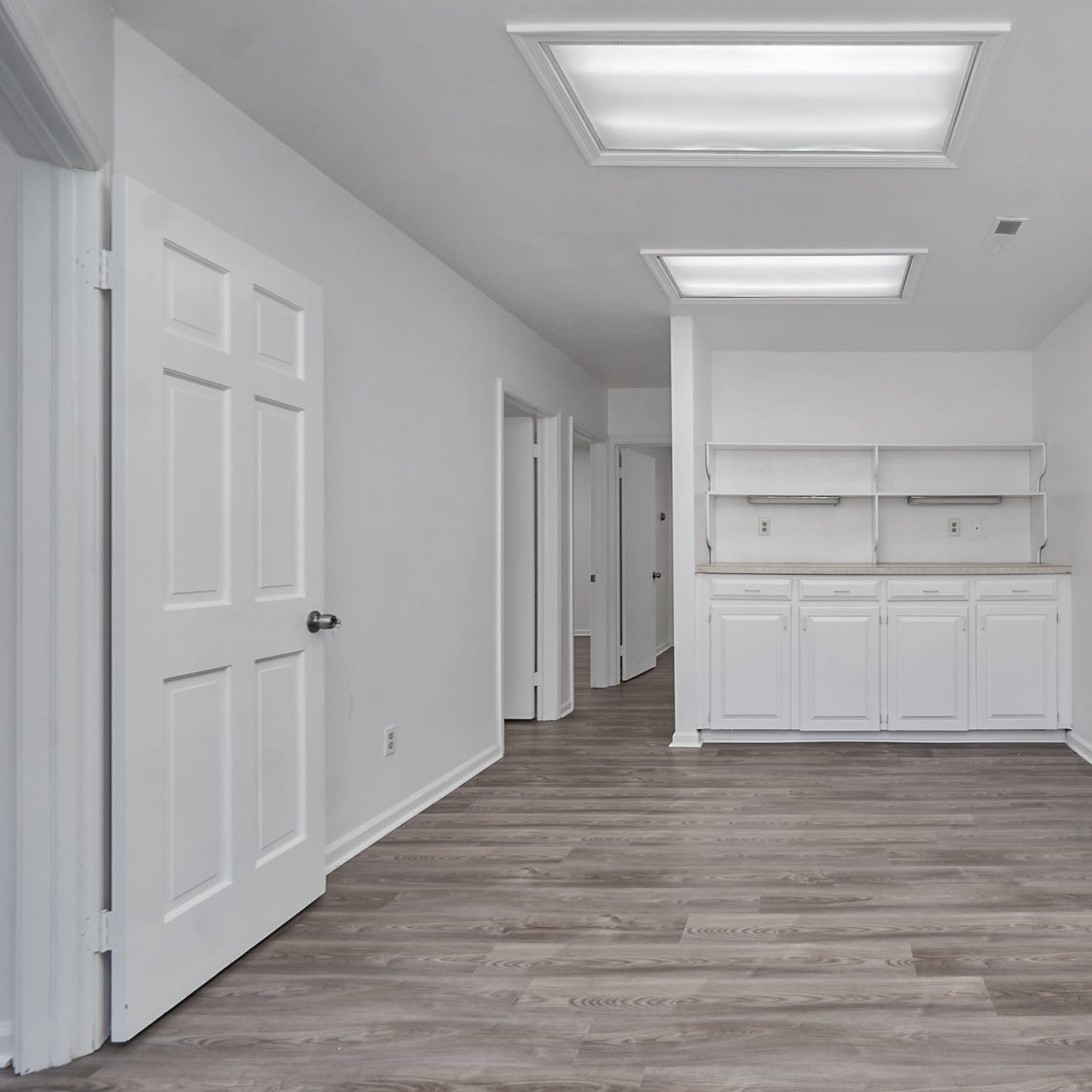 A spacious, empty room with light-colored walls, a wooden floor, and a row of white cabinets against one wall.