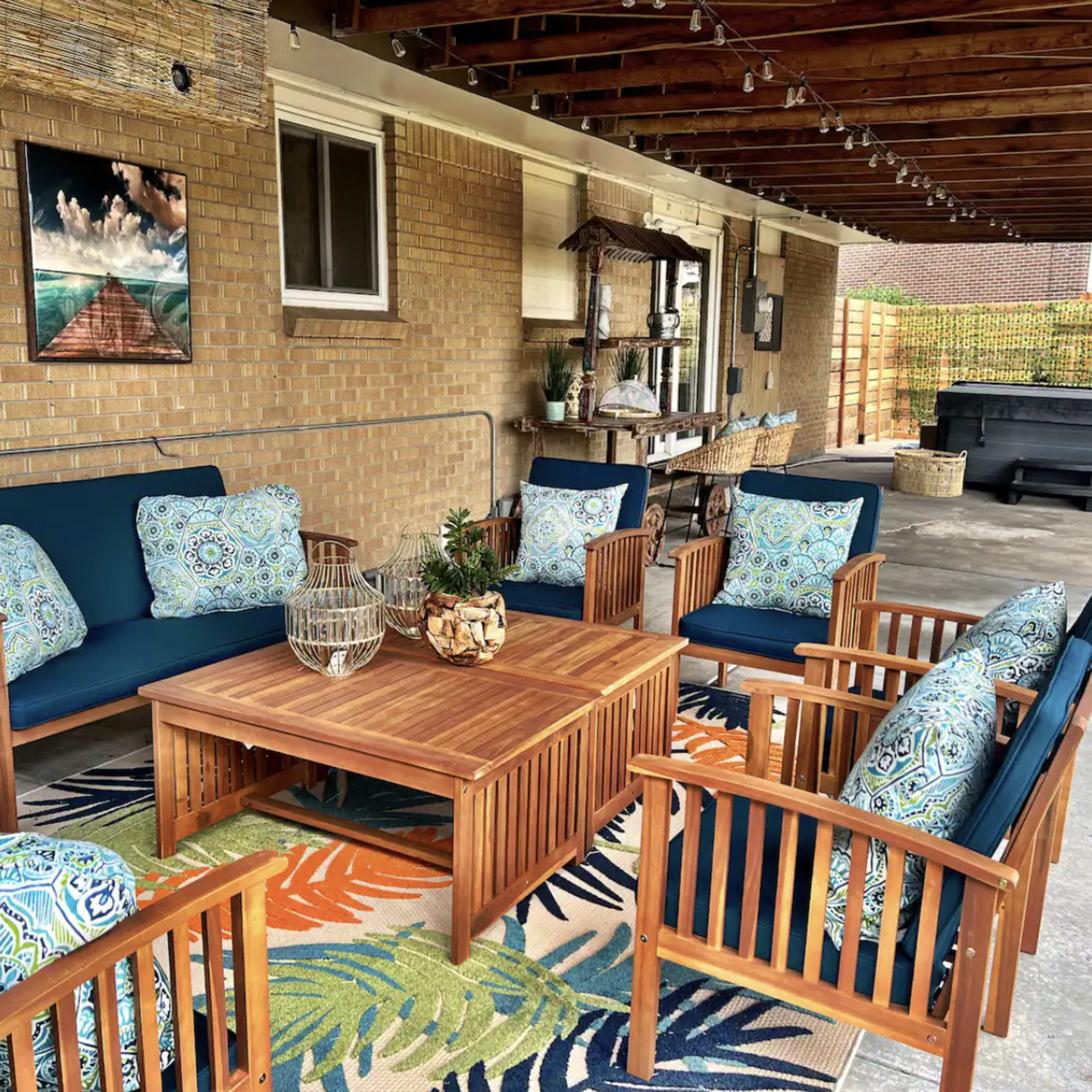 A covered outdoor patio area furnished with blue chairs, a wooden table, decorative cushions, and a patterned rug.
