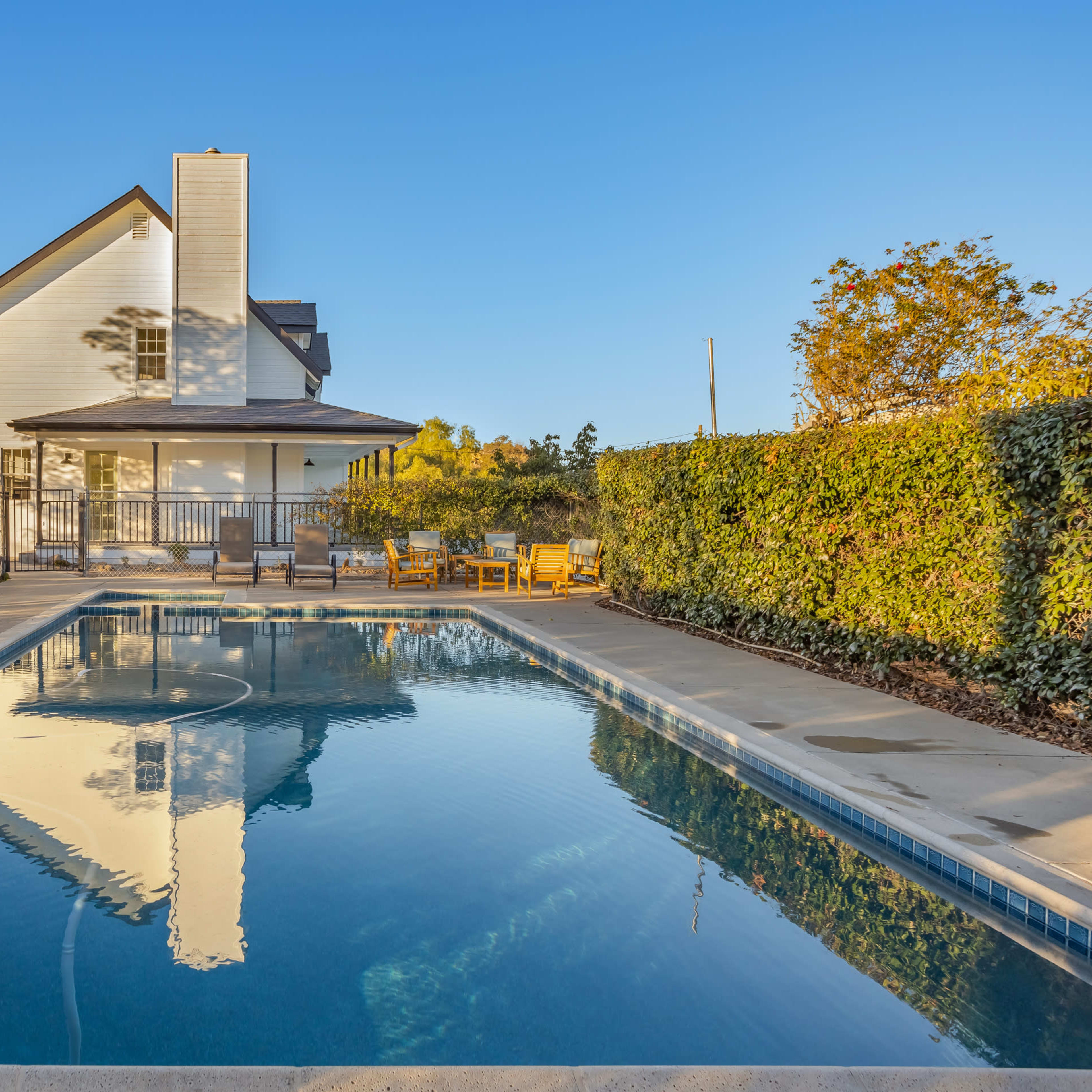 A clear blue swimming pool reflects a white house surrounded by green hedges and trees.