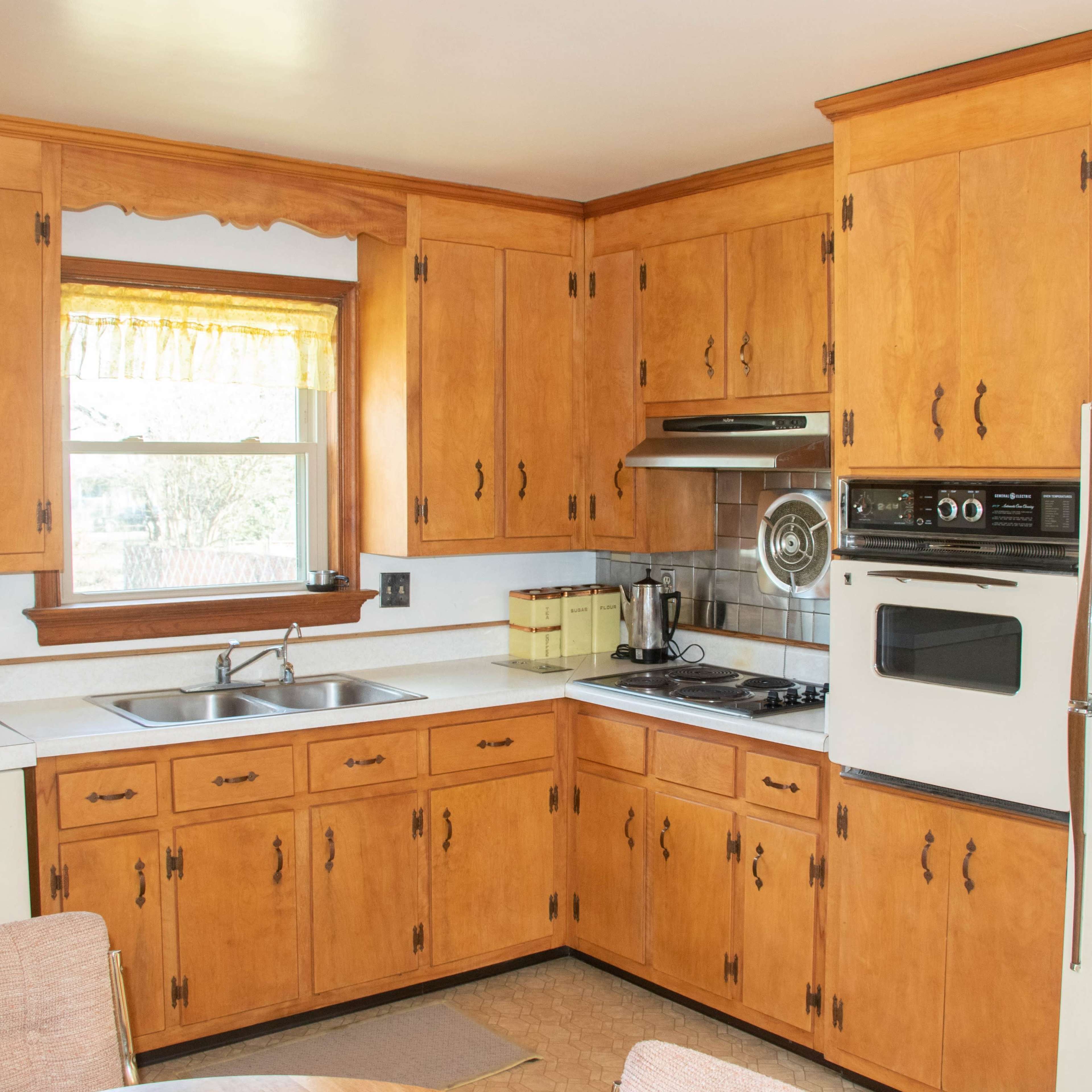 A kitchen with wooden cabinetry, a sink, a stove, and a white refrigerator, illuminated by natural light from a window.