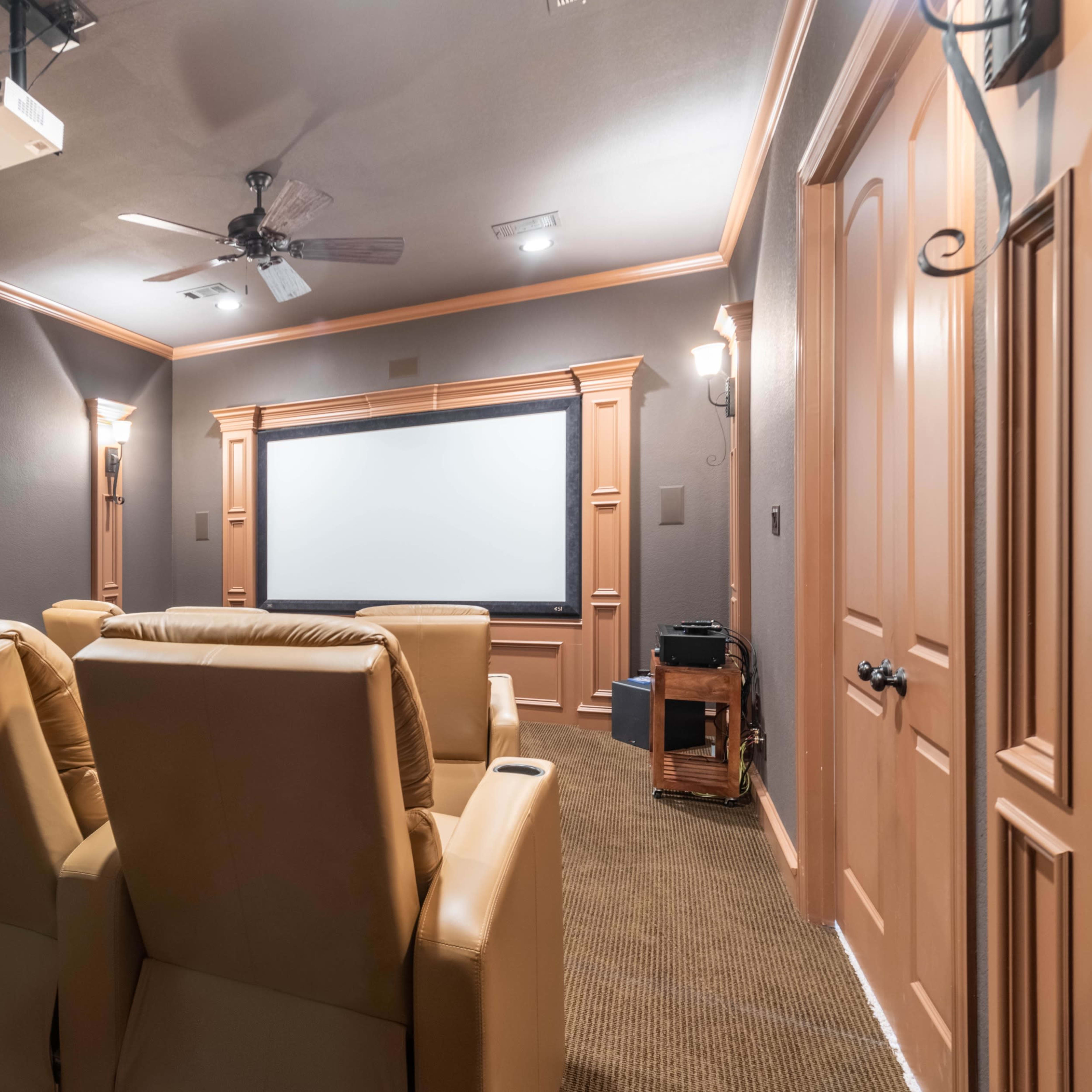 A home theater room featuring beige reclining chairs facing a large projection screen, with a projector mounted on the ceiling and wall sconces providing light.
