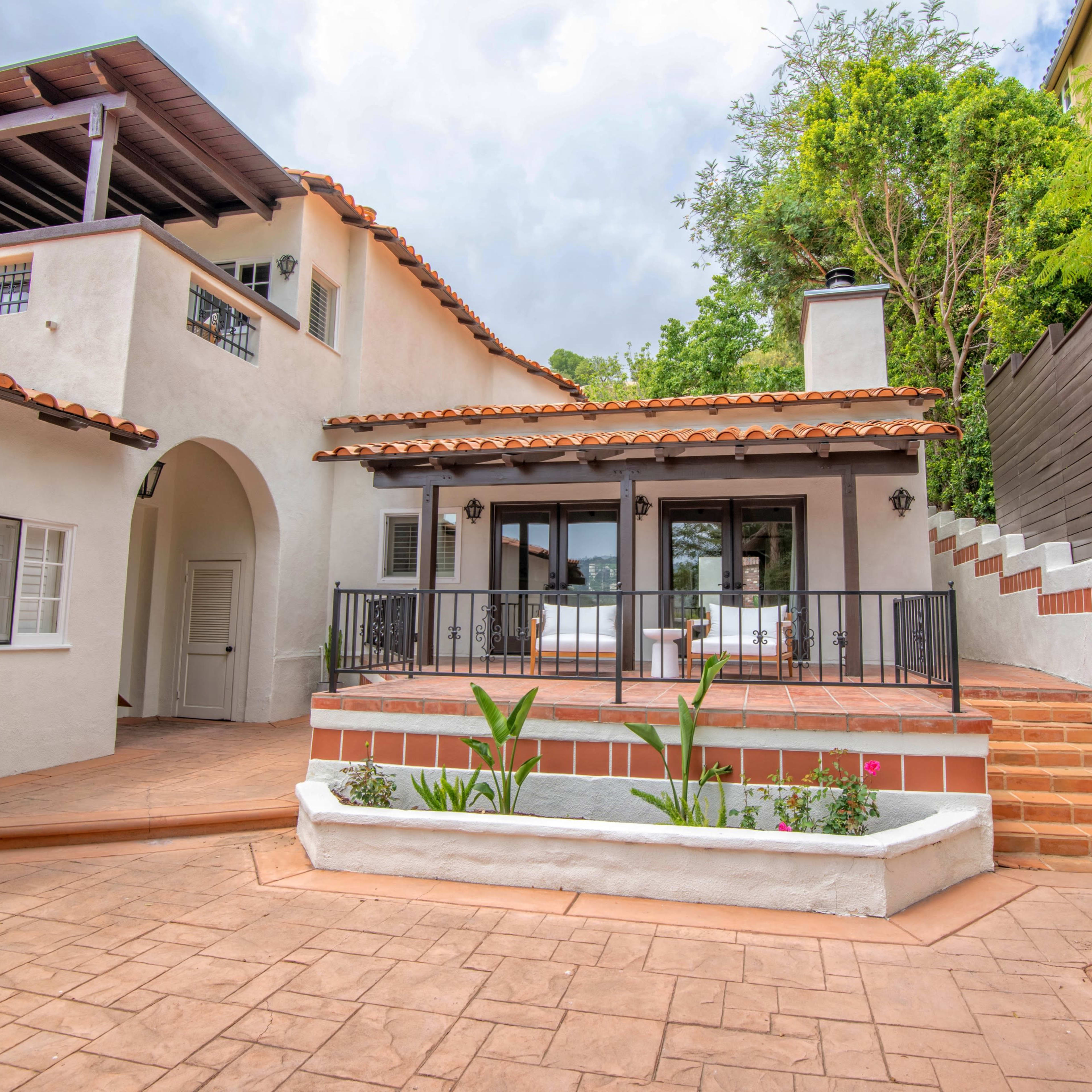 A two-story house with a terracotta roof, a covered balcony, and a landscaped outdoor terrace leading to a set of stairs.