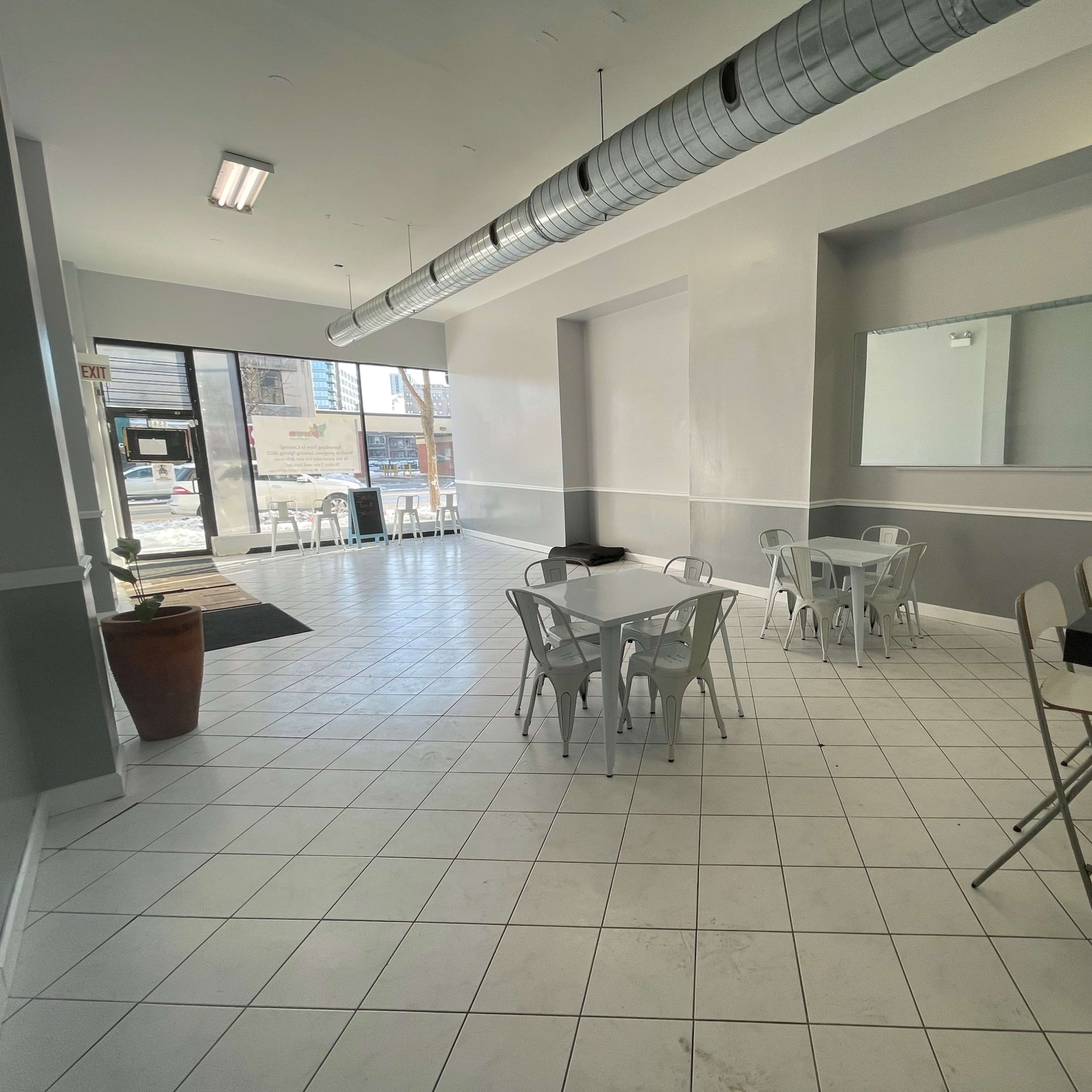 A modern, empty café with white walls, tiled flooring, and metal tables and chairs, featuring a large window at the entrance.