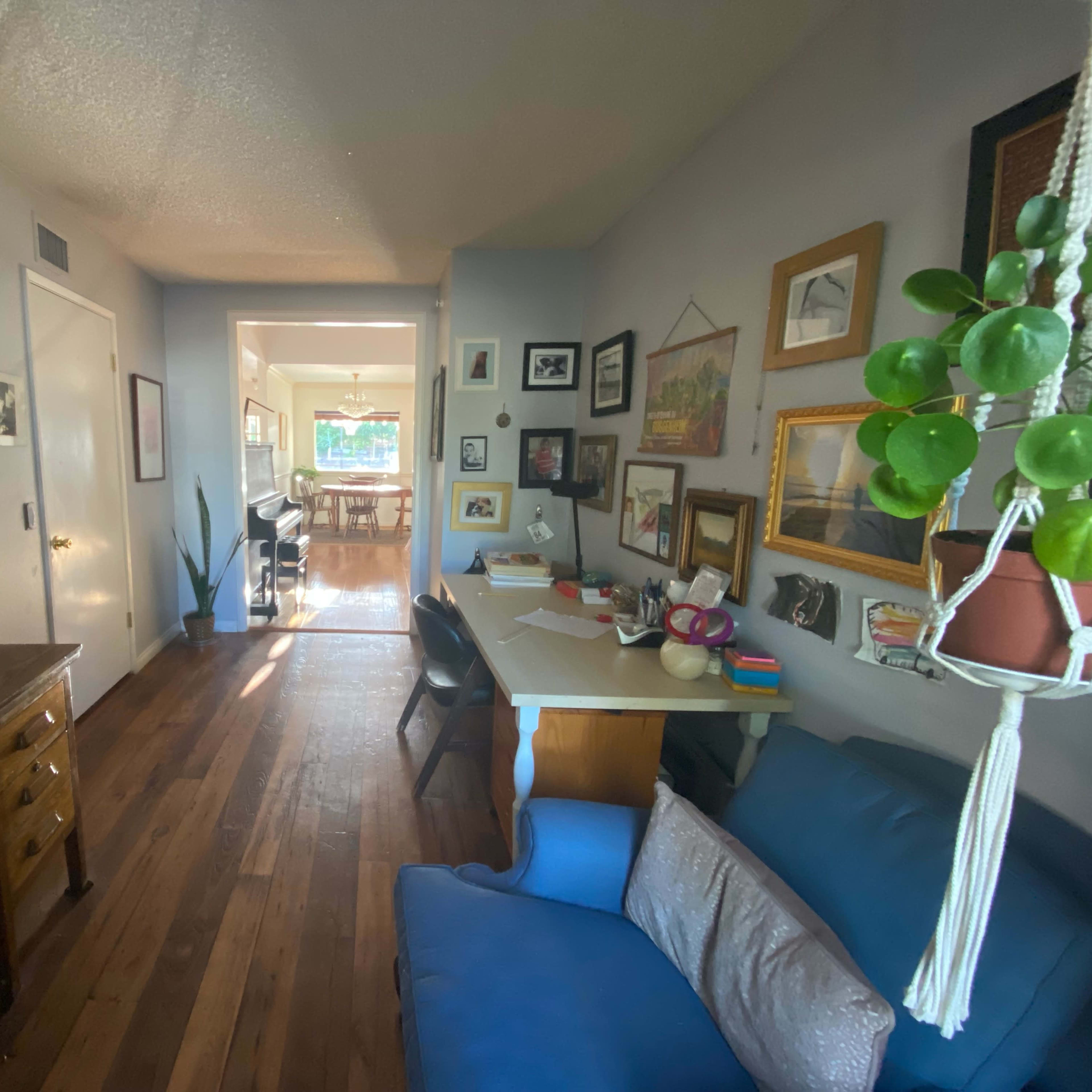 A hallway leads from a small desk area to a well-lit dining space, with framed art on the walls and a blue sofa nearby.