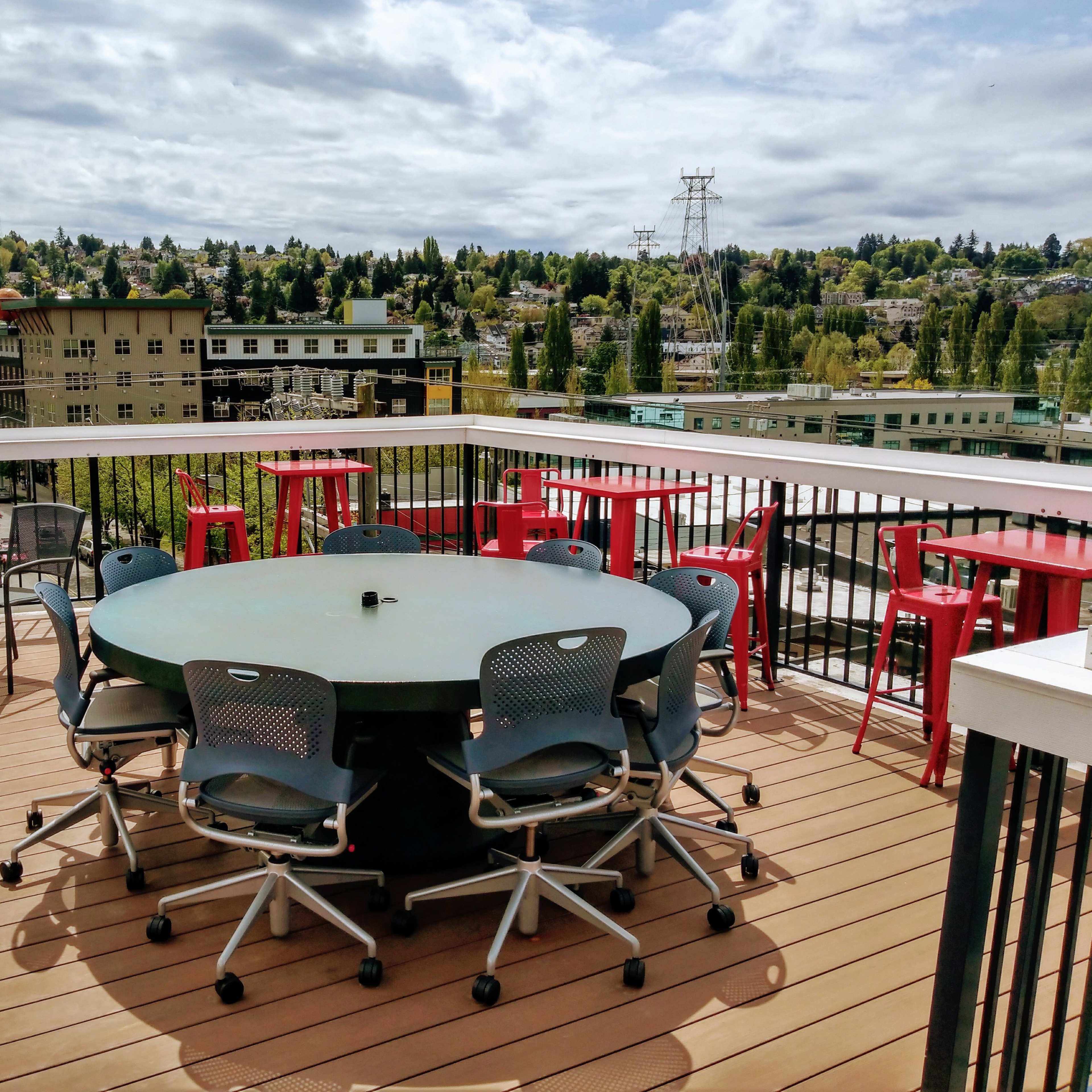 A rooftop terrace with a round table surrounded by chairs, a red umbrella, and a view of buildings and trees in the background.