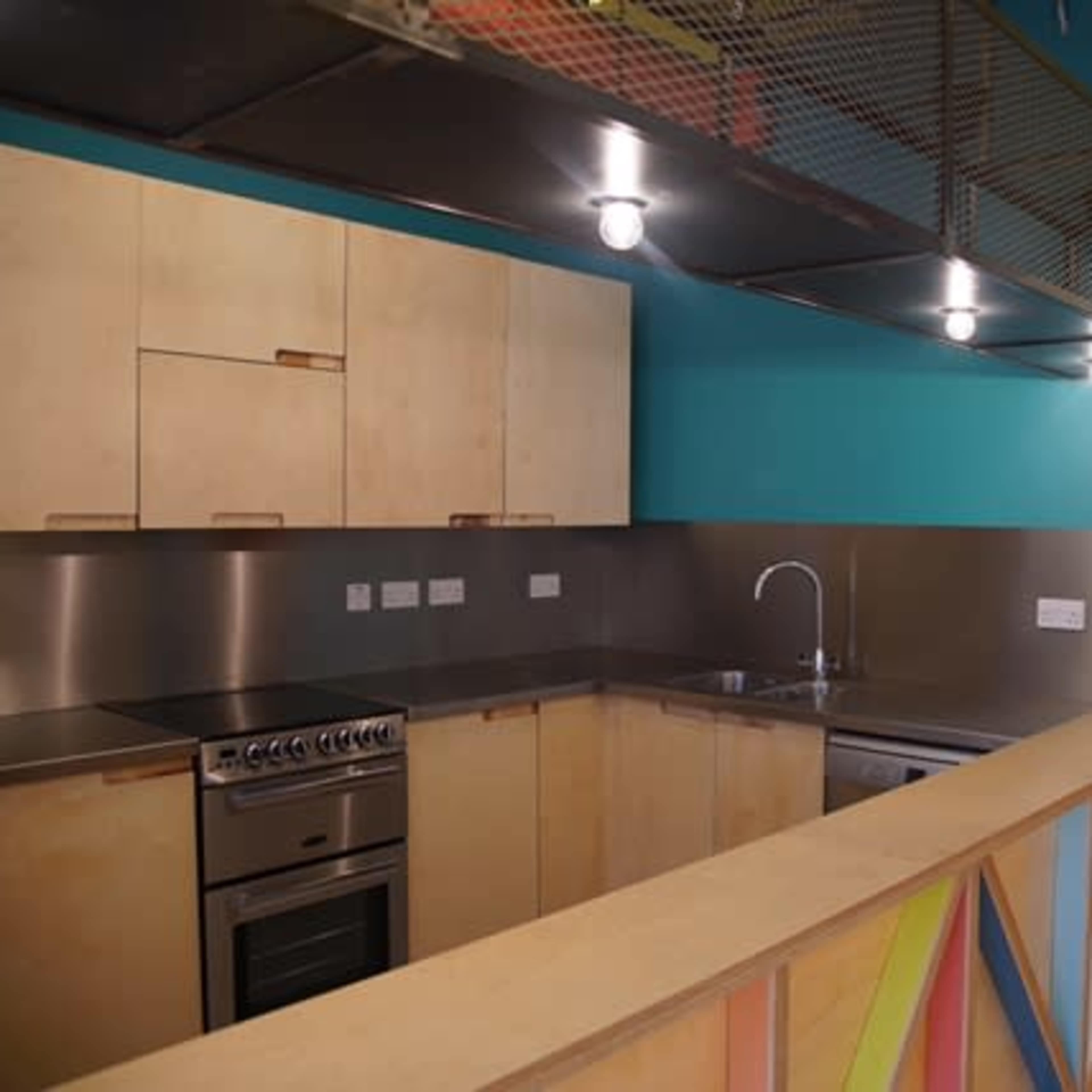 A modern kitchen with wooden cabinetry, stainless steel appliances, and a bright blue wall, with a view of a hallway beyond.