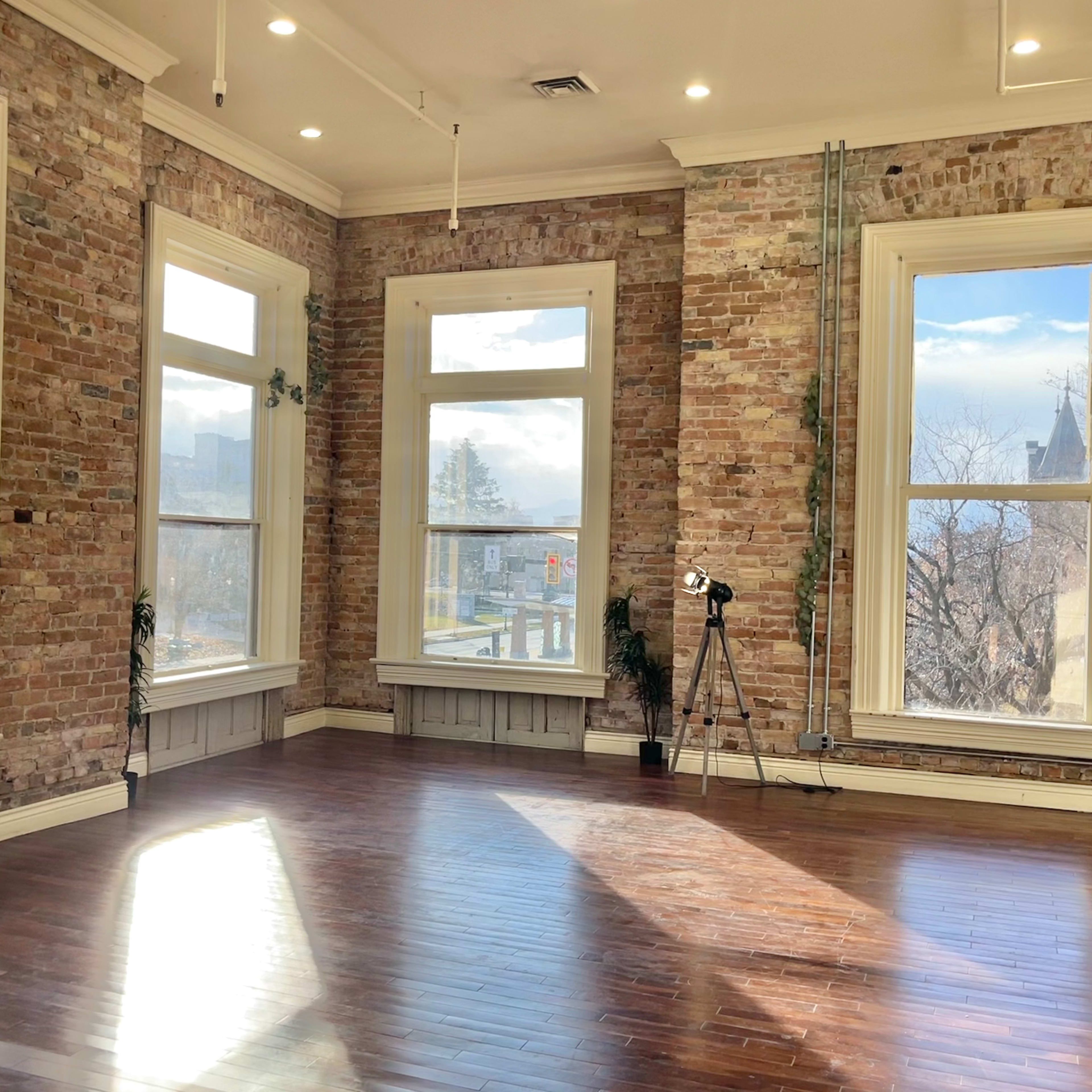 A spacious, well-lit room with exposed brick walls, large windows, and polished wooden floors.