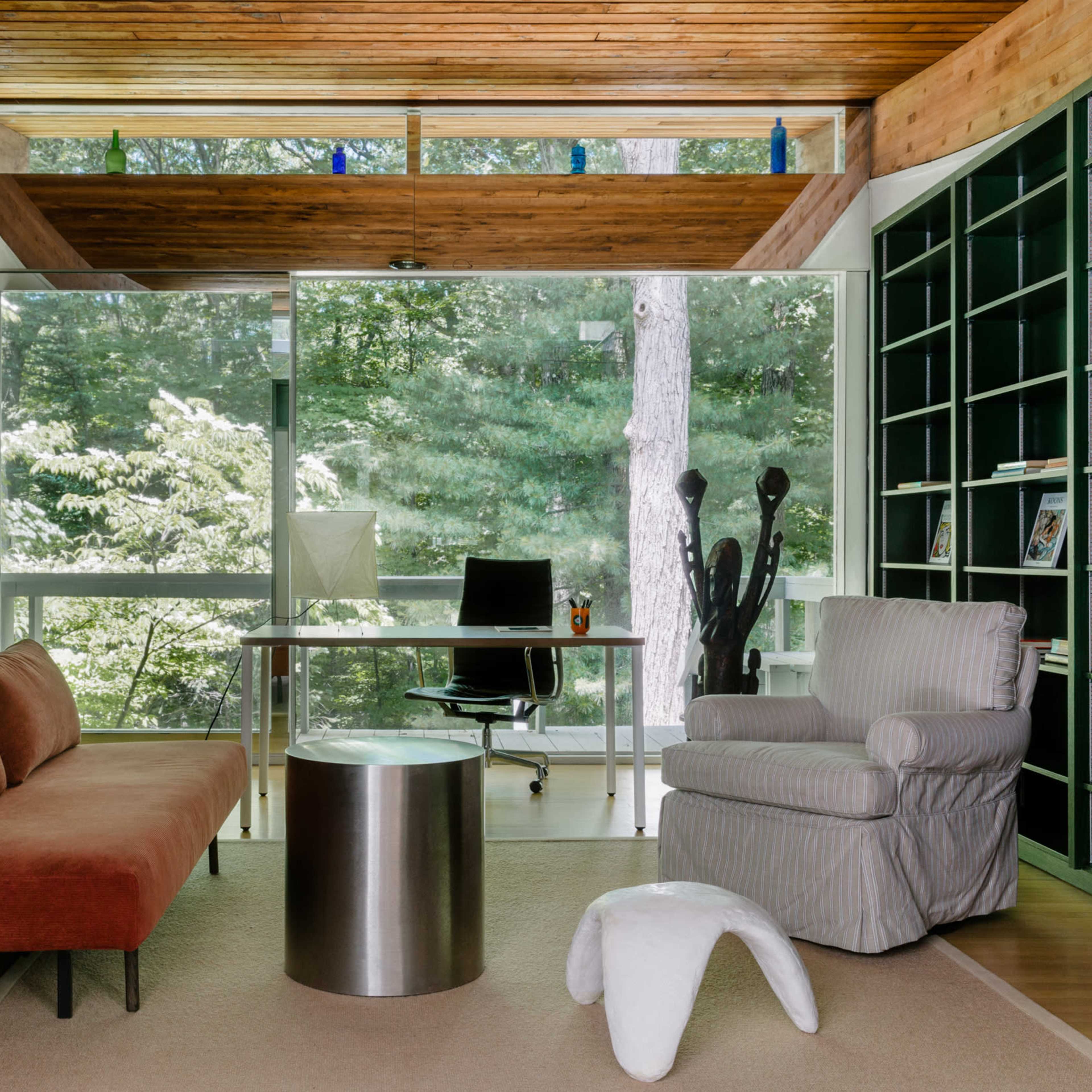 A modern home office featuring a brown sofa, a silver cylindrical coffee table, a white plush ottoman, a desk with a computer, and large windows overlooking a forested view.