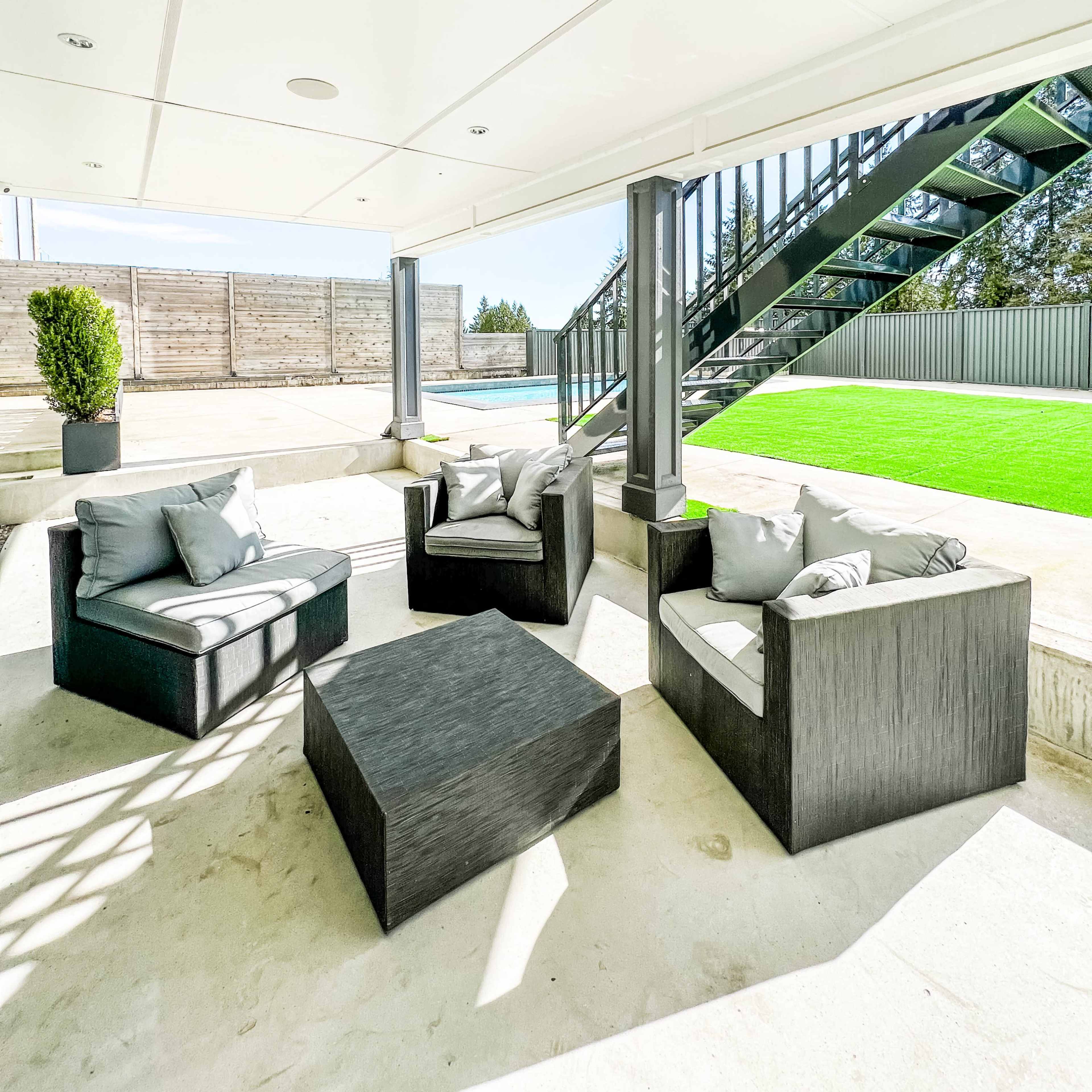 A modern outdoor seating area with black furniture arranged around a square table, set beneath a covered patio near a pool area.