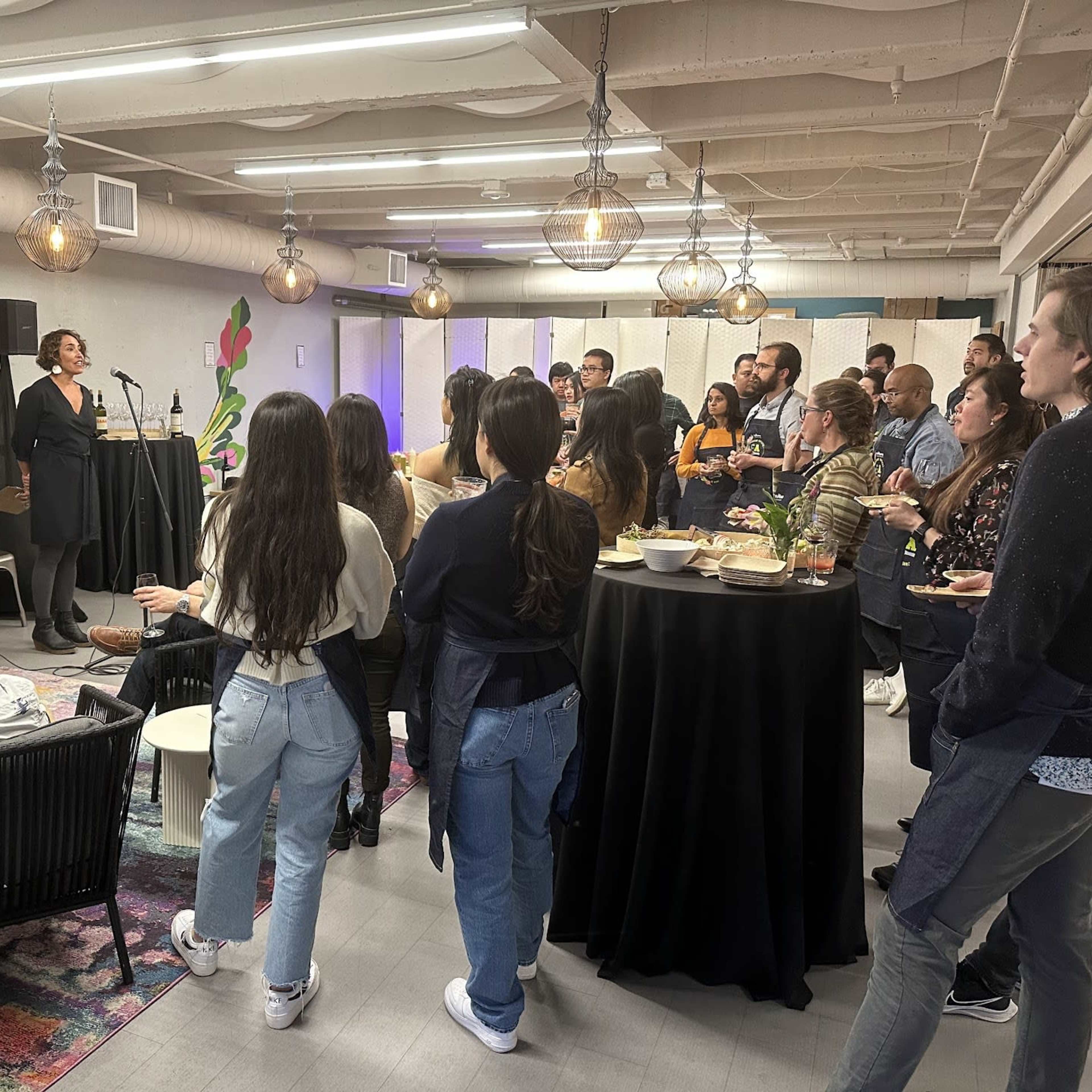 A group of people attentively listens to a speaker at a social event in a modern indoor space with decorative lighting and a colorful mural.