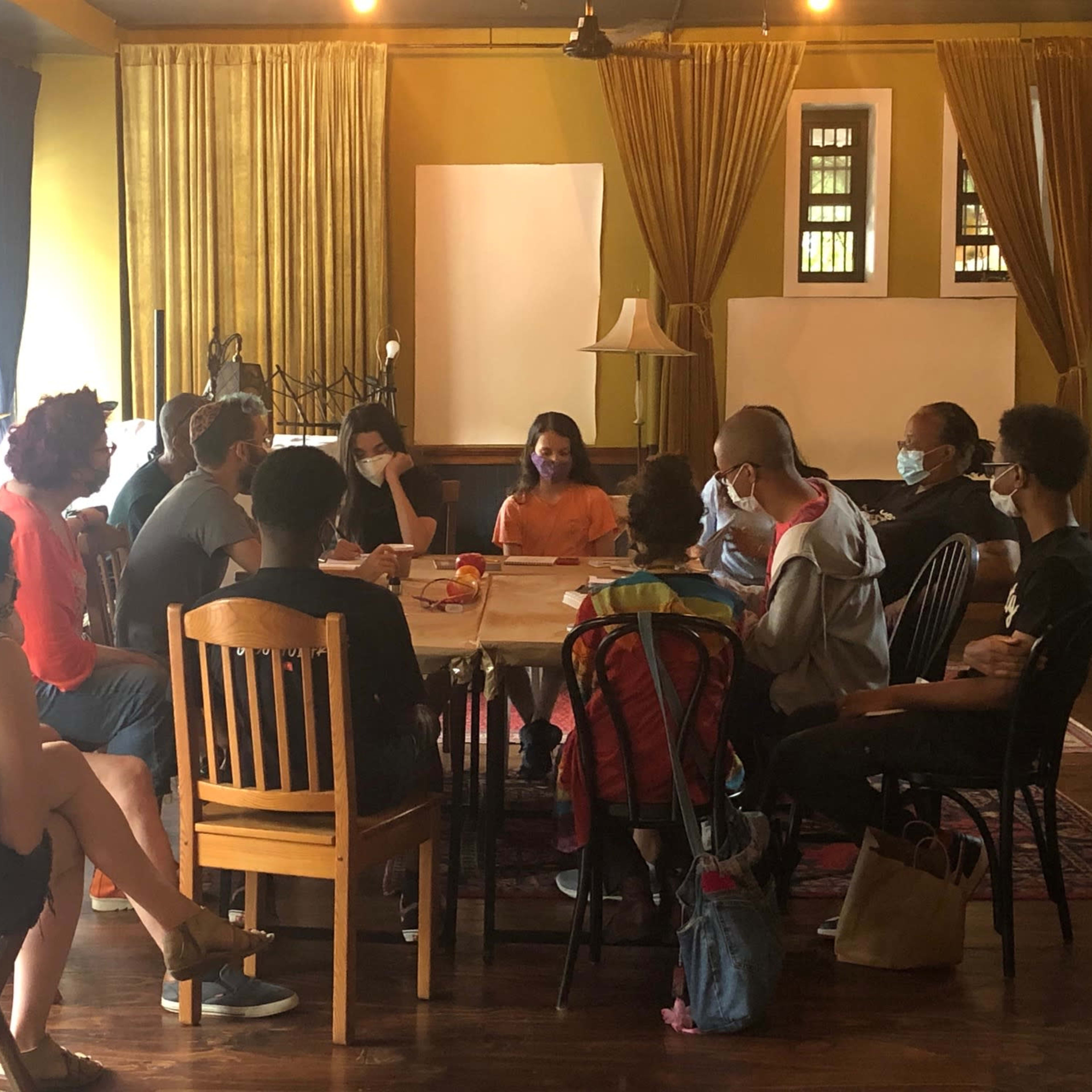 A group of people wearing masks is seated around a table in a room with yellow walls and curtains.