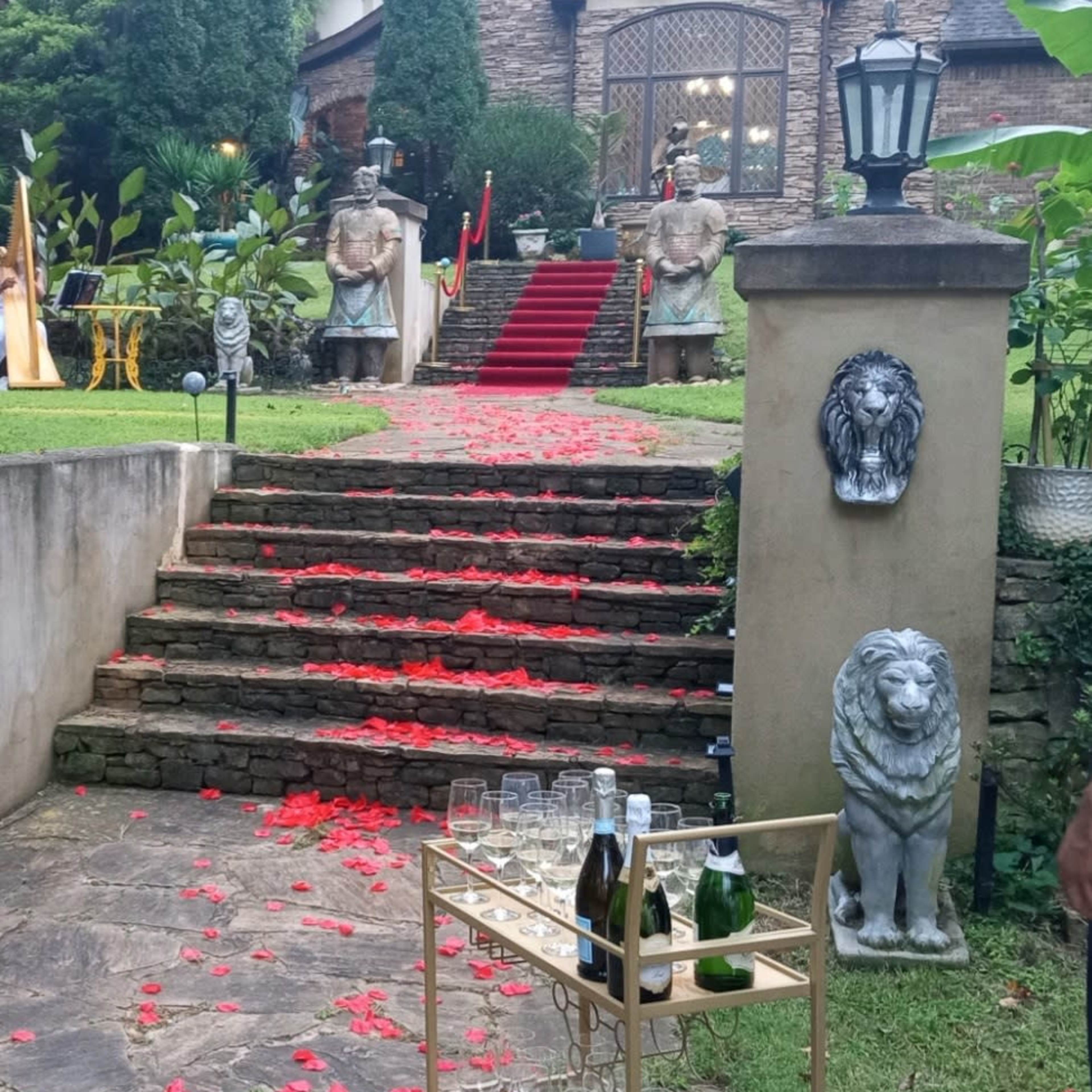 A stone pathway lined with red rose petals leads to a grand house featuring large statues and a lantern, with a cart of glasses and champagne nearby.