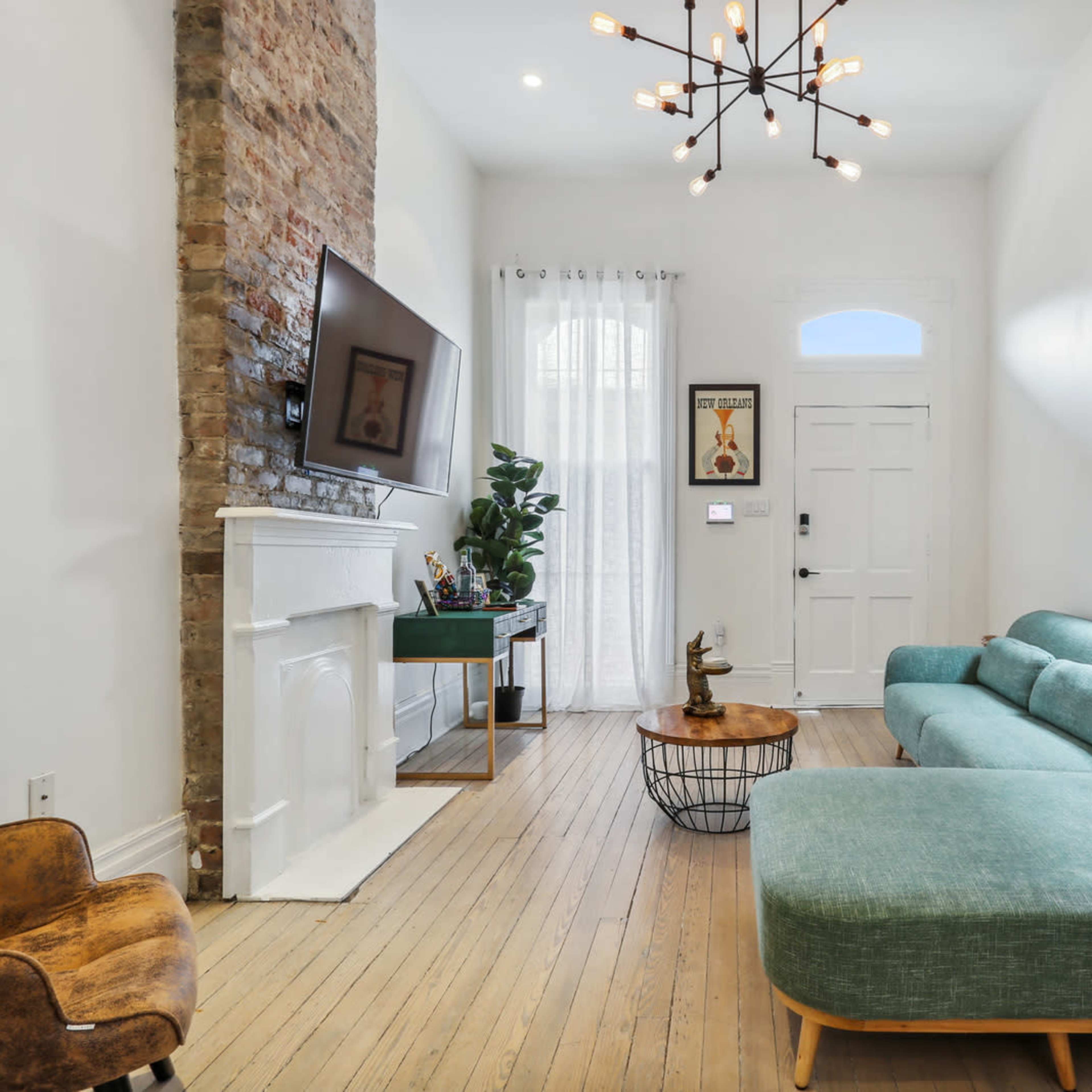 A living room with a green sectional sofa, a wooden coffee table, and a television mounted on a brick wall, illuminated by a modern chandelier.
