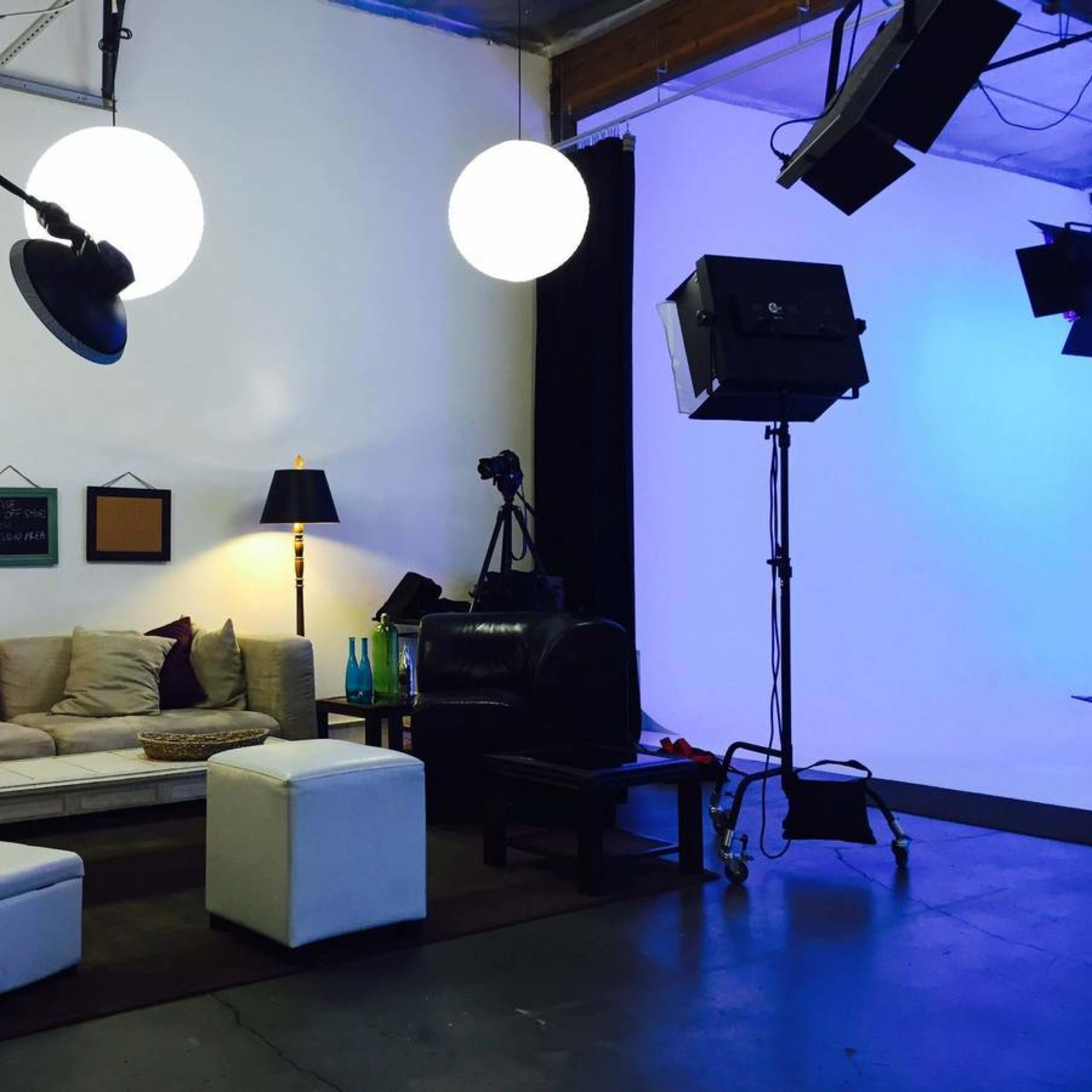 A modern studio setup with a beige couch, white ottomans, a floor lamp, and photography equipment against a backdrop of a blue-lit wall.