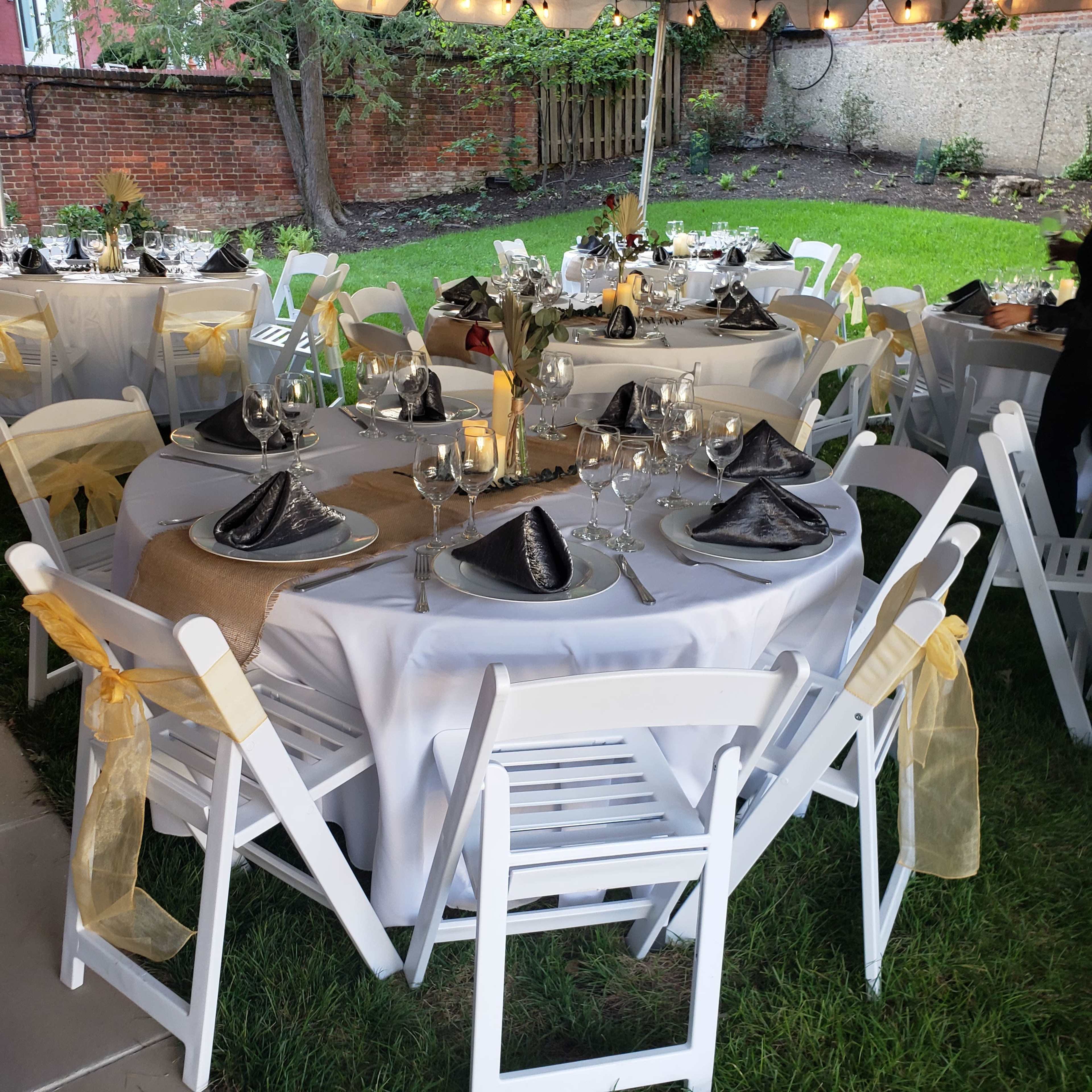 A decorated outdoor dining setup features round tables with white tablecloths, black napkins, and elegant glassware, surrounded by green grass and string lights overhead.