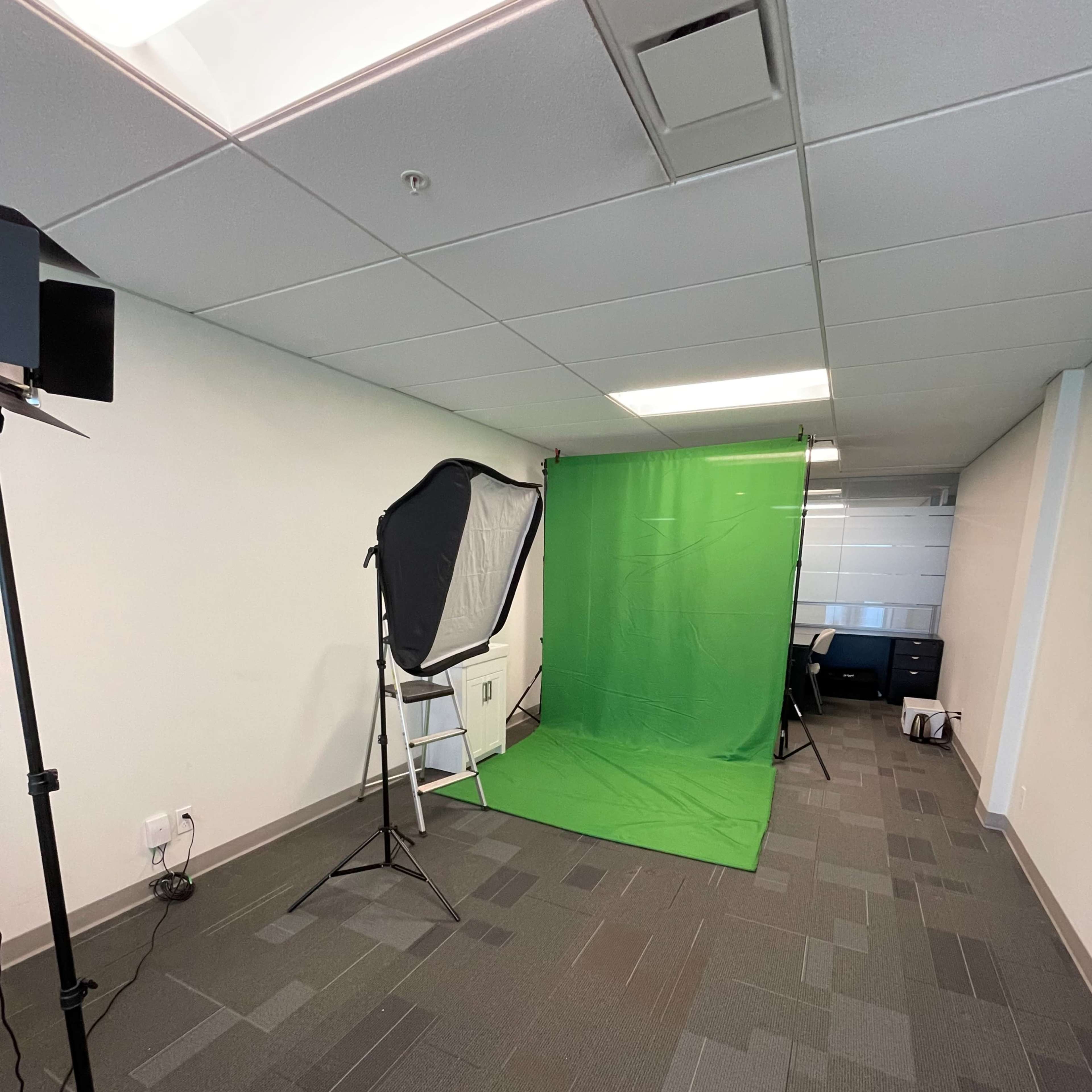 An empty office space with a green screen setup, a softbox light, and a camera stand.