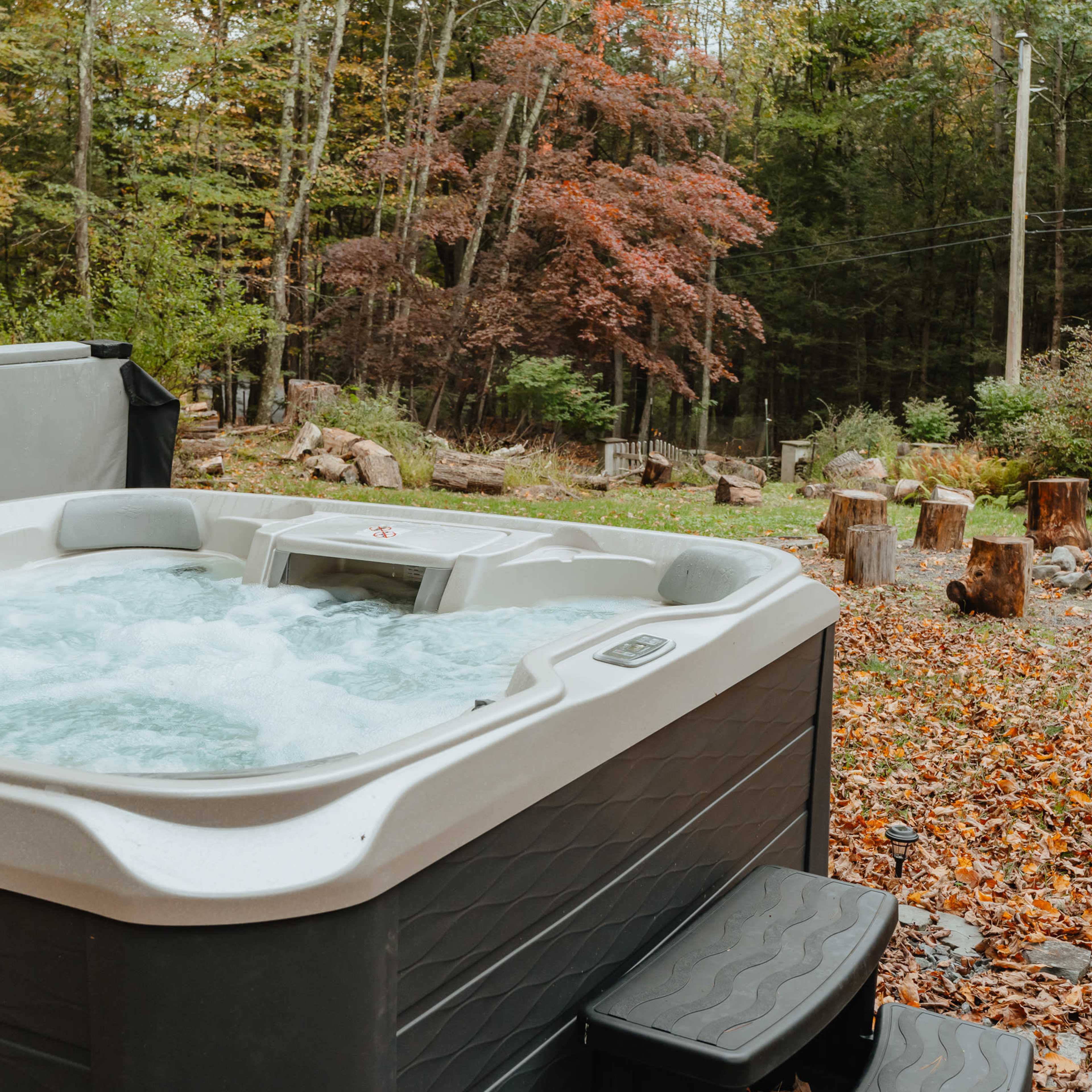 A hot tub is situated in a yard surrounded by trees with autumn foliage and a fire pit nearby, amidst scattered leaves.