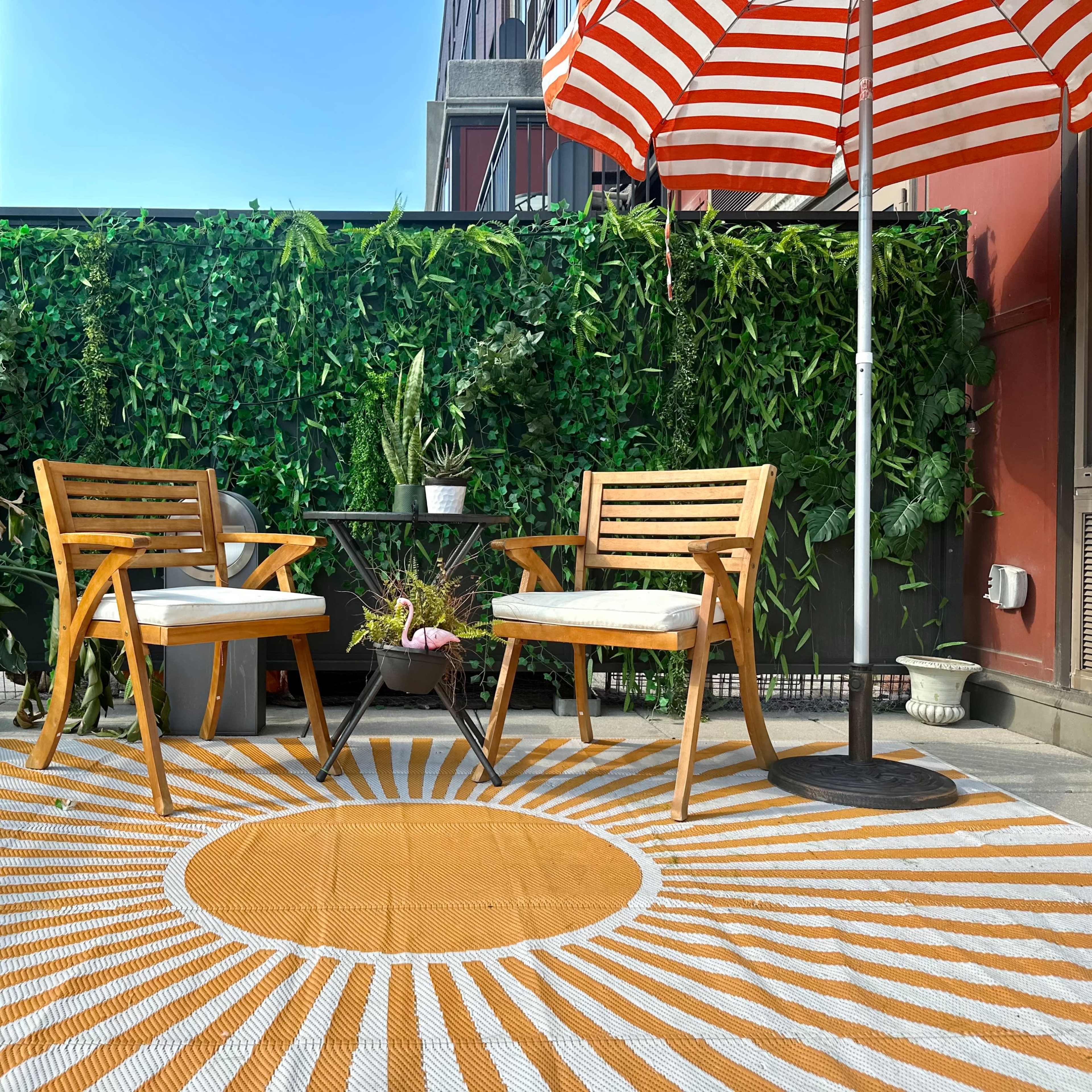 A balcony with two wooden chairs, a small round table, and a sun-patterned rug, surrounded by greenery and an umbrella.