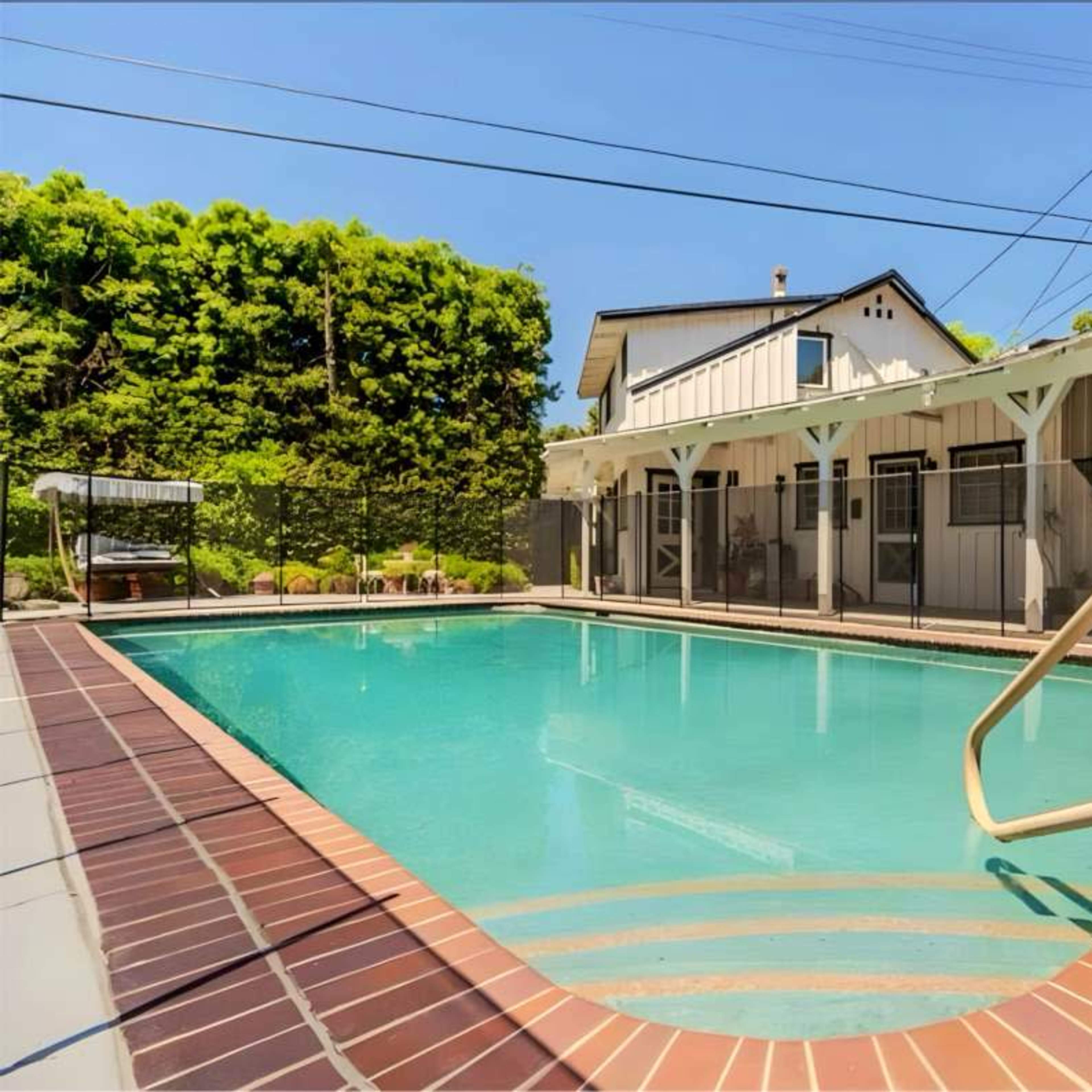 A rectangular swimming pool is surrounded by a tiled deck and is located next to a house with several adjacent patio spaces.