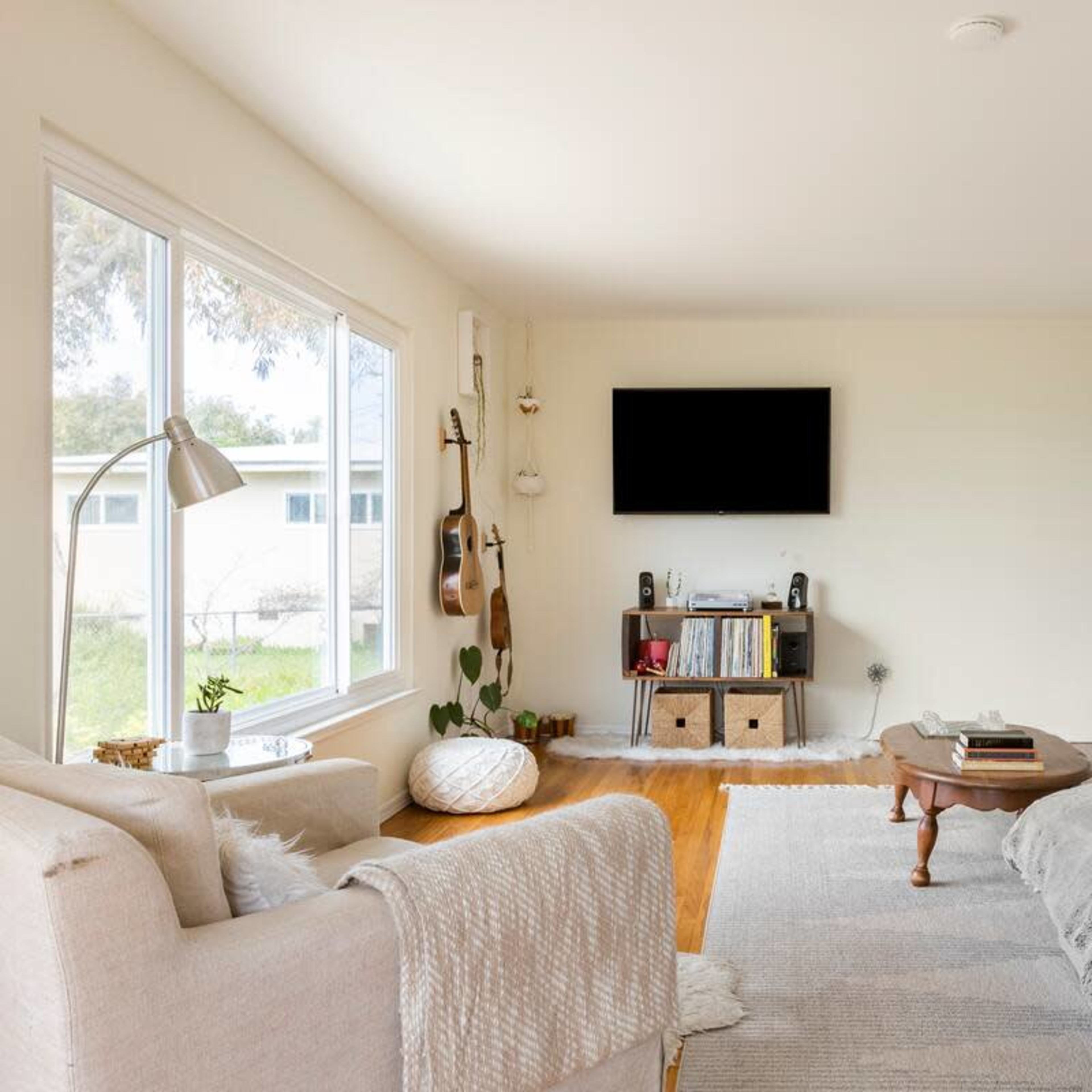 A light-filled living room features a gray sofa, a plush armchair, and a wooden coffee table, with a wall-mounted TV and shelves displaying books and decor.