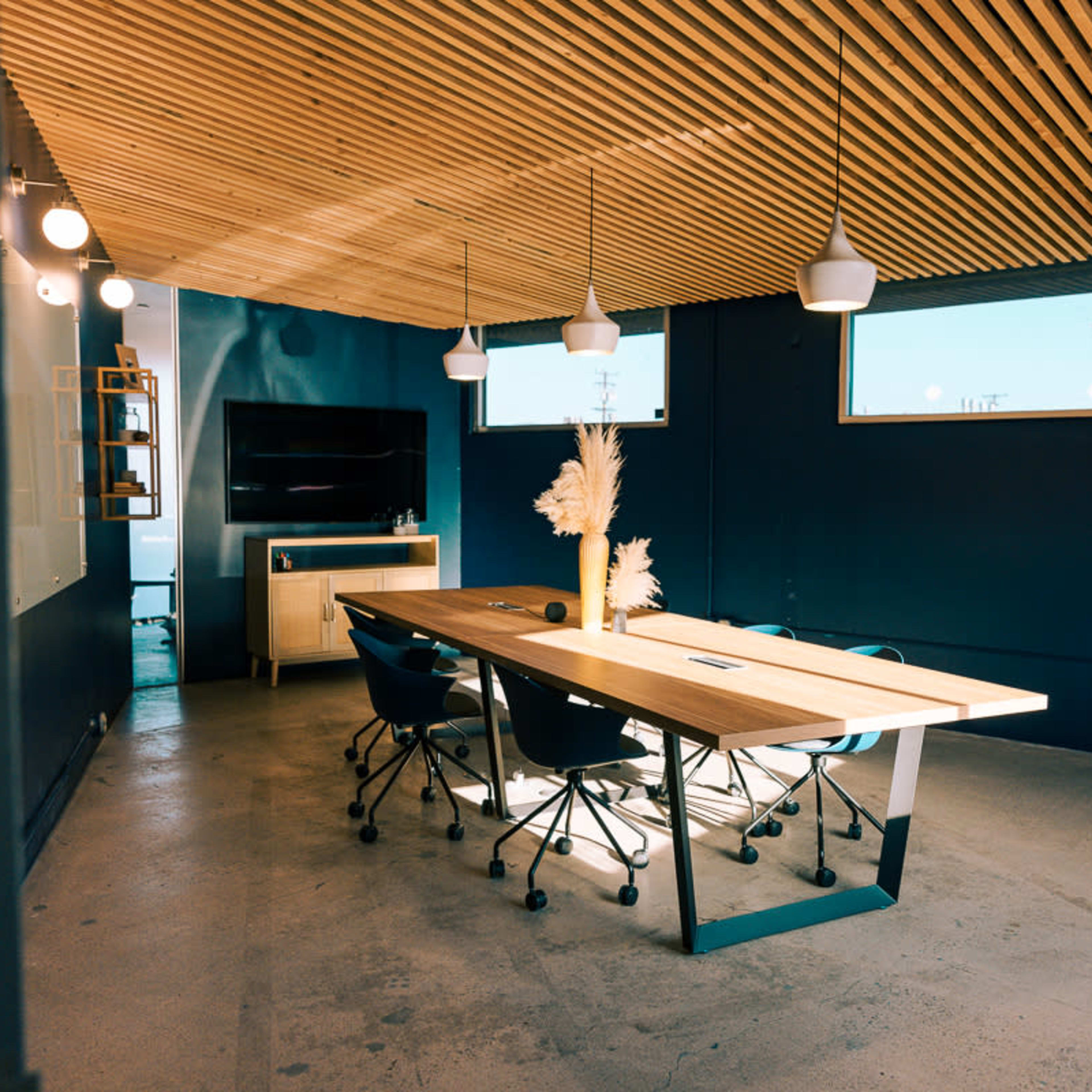 A modern conference room featuring a large wooden table surrounded by rolling chairs, with a plant and a wall-mounted TV in the background.