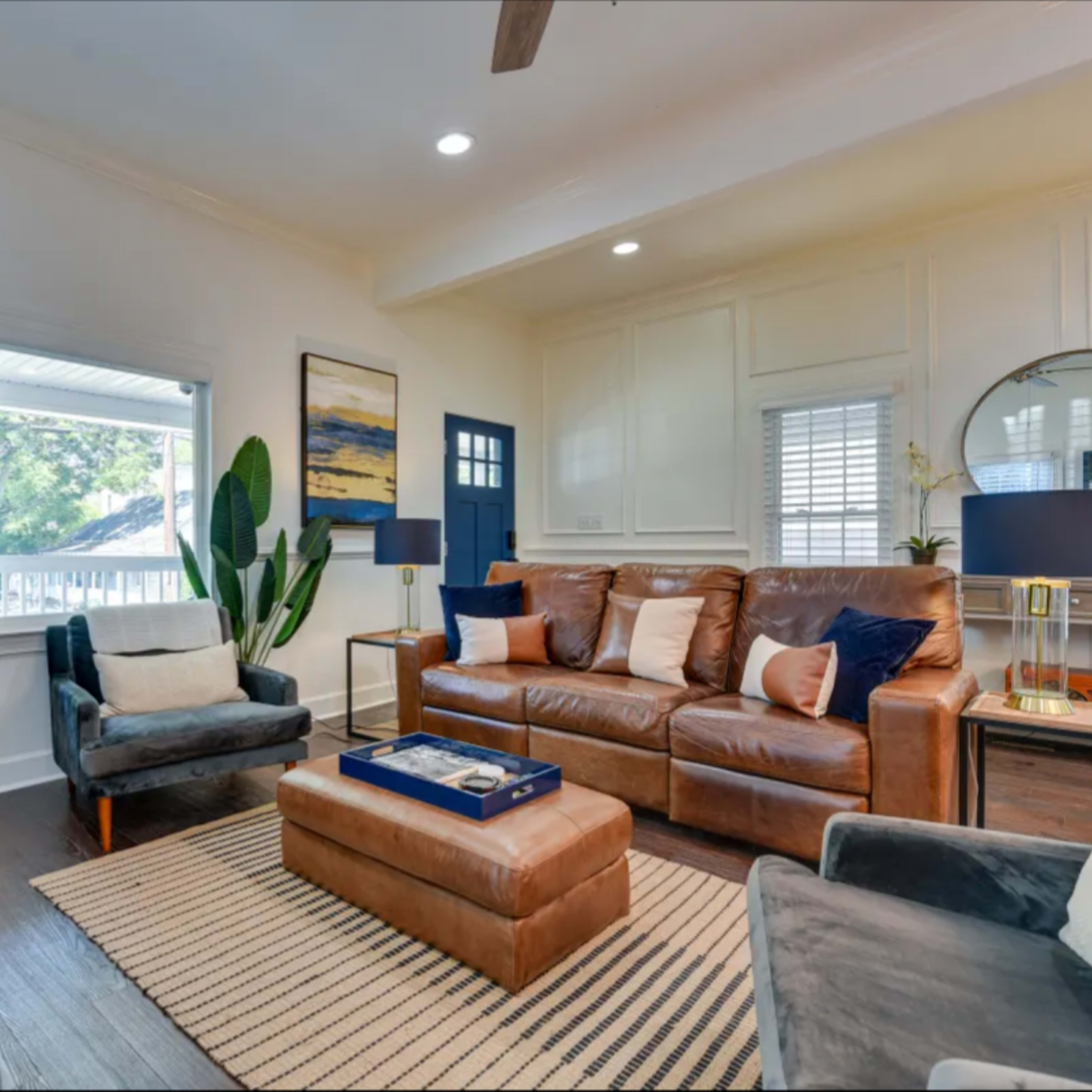 The living room features a leather sofa, an armchair, a coffee table, a wall-mounted TV, and a potted plant, with ample natural light coming through the windows.