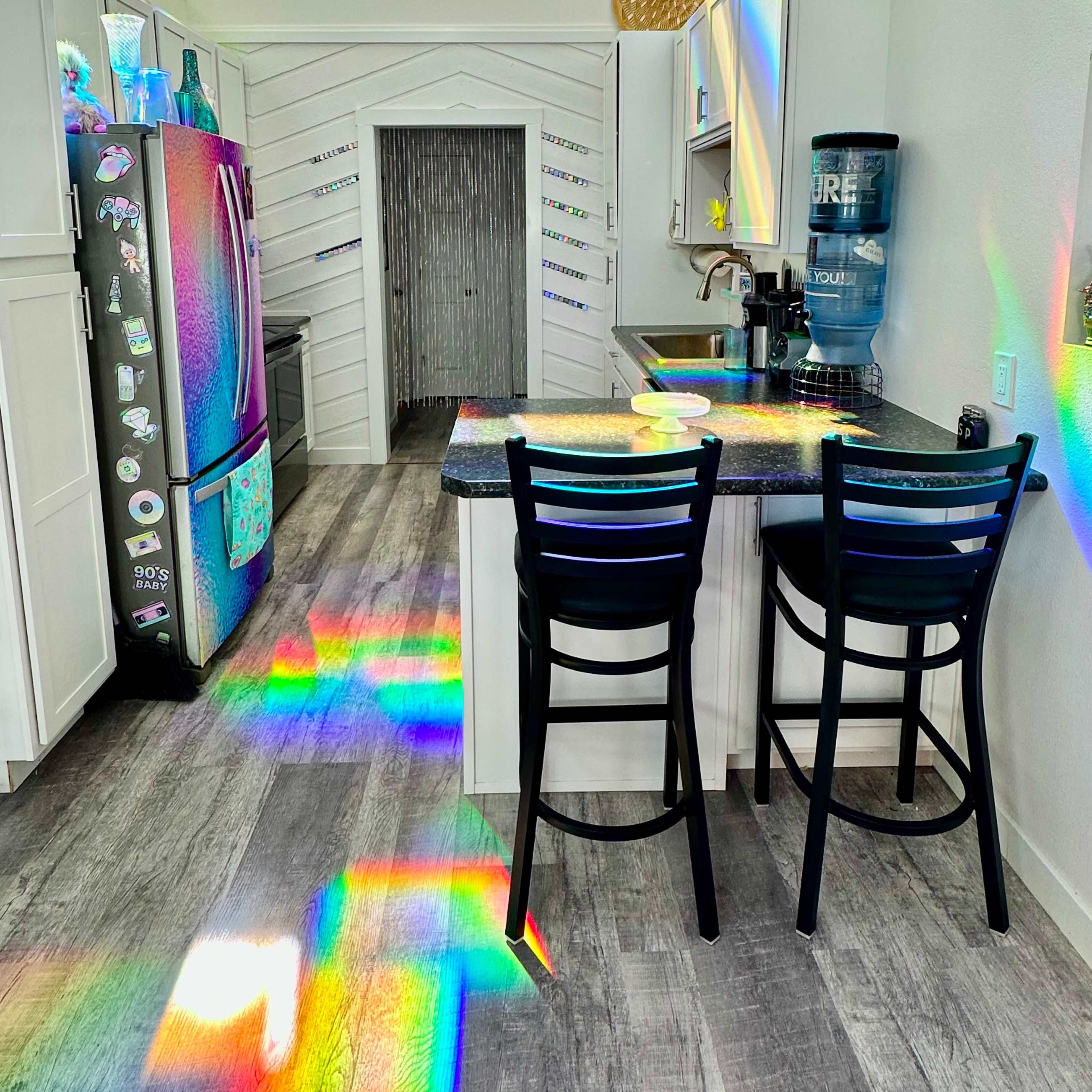 A bright kitchen features a refrigerator, a water dispenser, and two bar stools, with sunlight creating rainbow reflections on the countertop.
