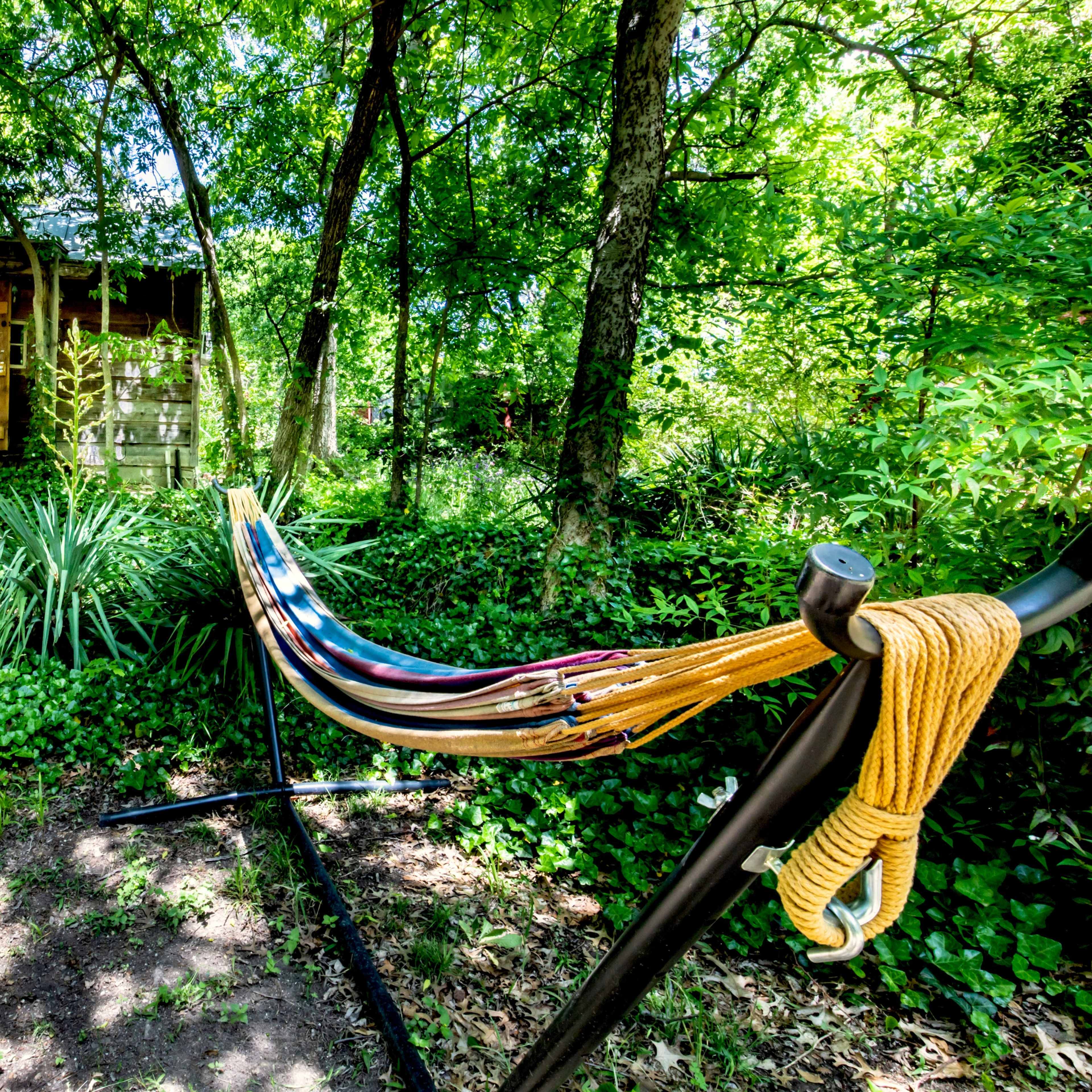 A colorful hammock is hung beside a path leading to a small cabin surrounded by lush greenery.