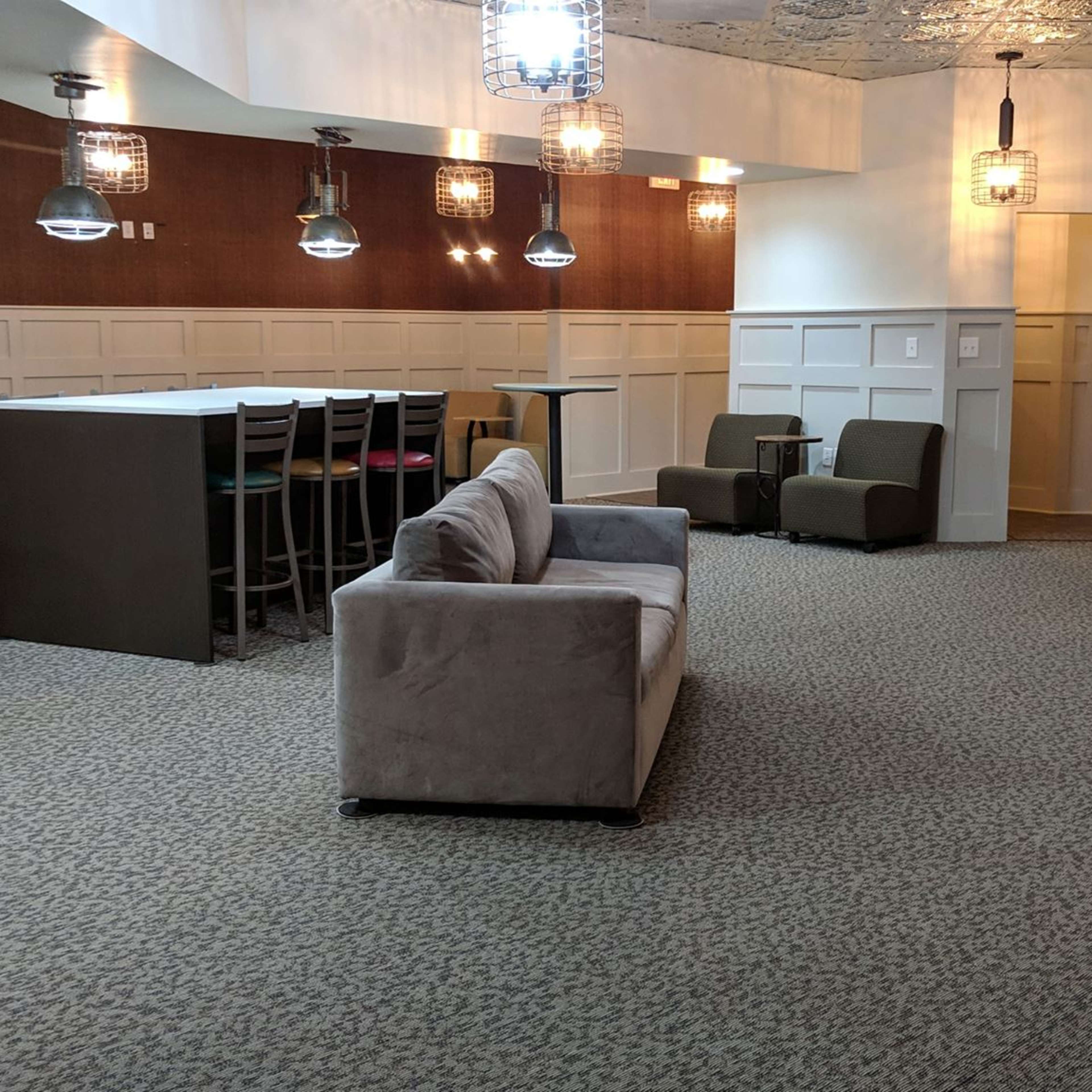 A modern lounge area featuring a gray sofa, two green chairs, a counter with high chairs, and pendant lighting against a paneled wall.