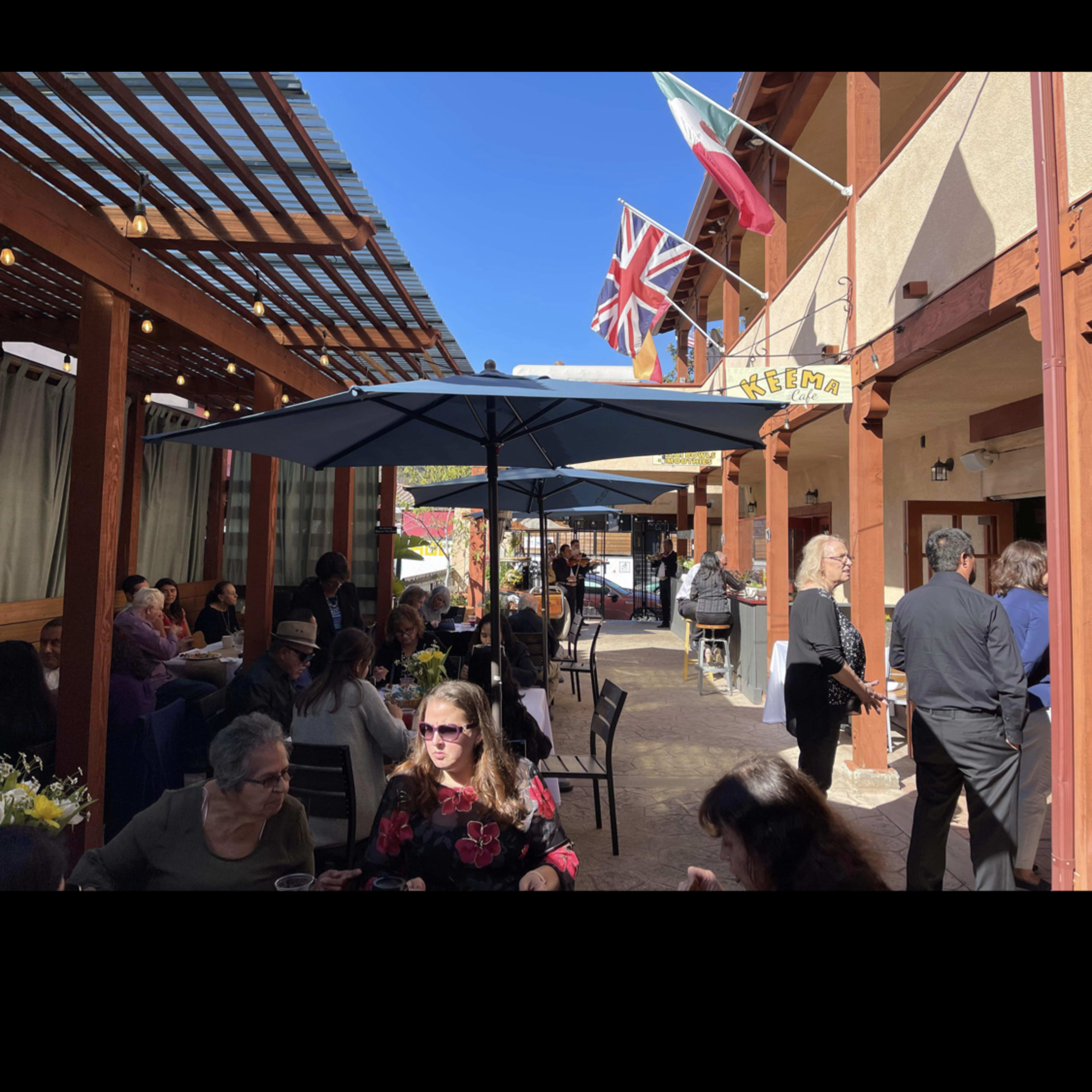 A lively outdoor dining area with people seated at tables under a canopy, while flags from various countries hang overhead.