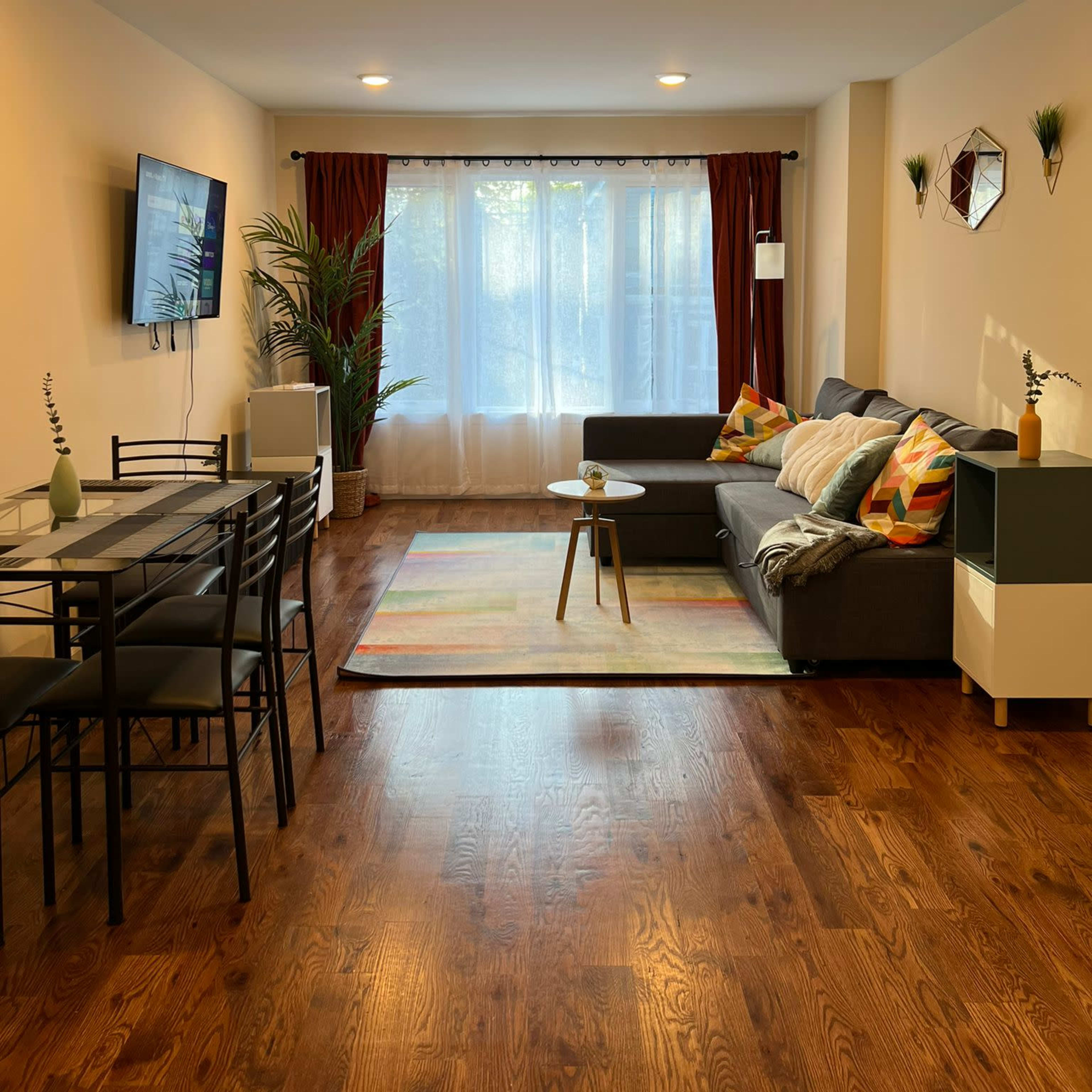 A modern living room features a gray sofa, a dining table with chairs, and large windows draped with curtains, allowing natural light to fill the space.
