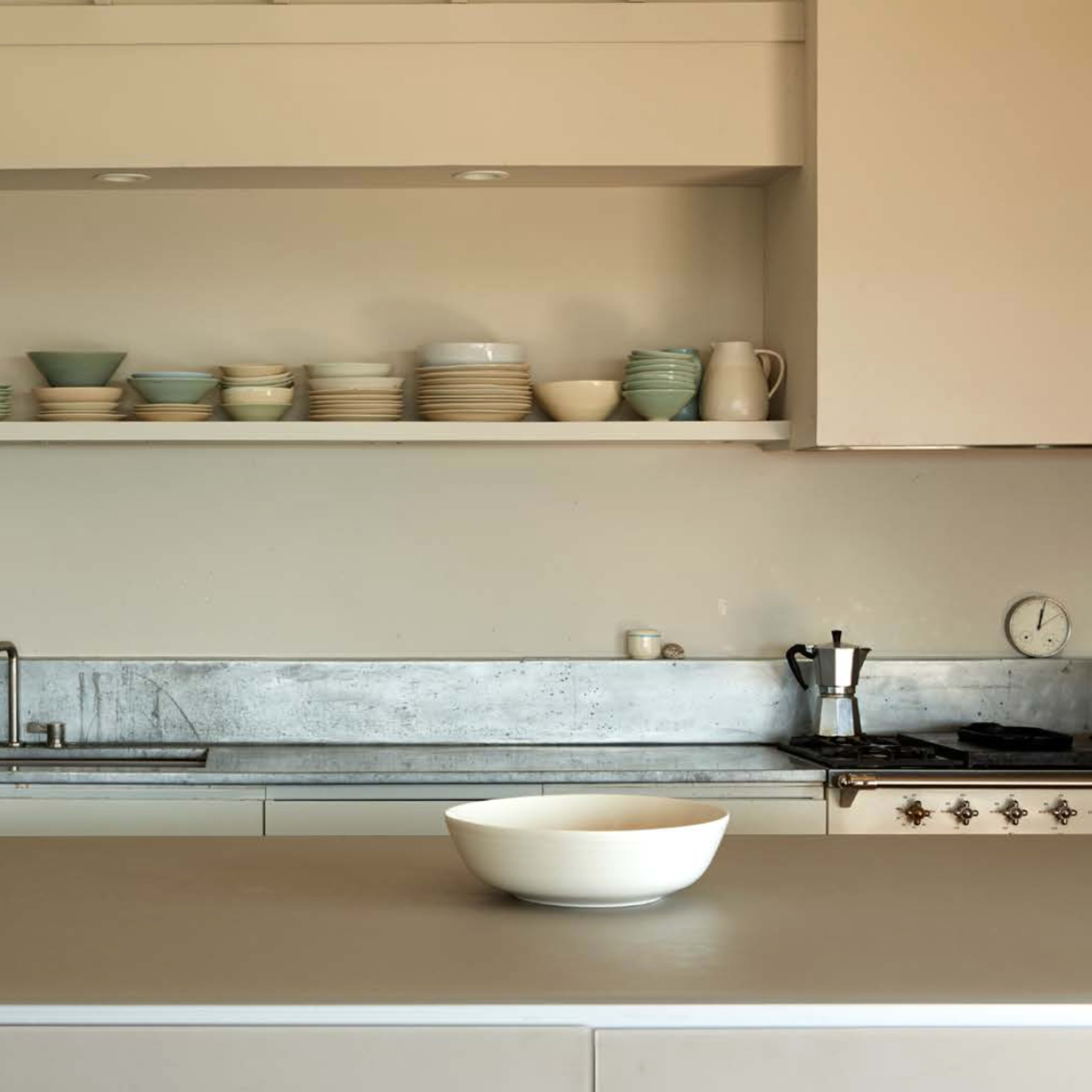 A modern kitchen with a minimalist design, featuring a large central island, shelves of dishware above, and a stainless steel sink and countertop.
