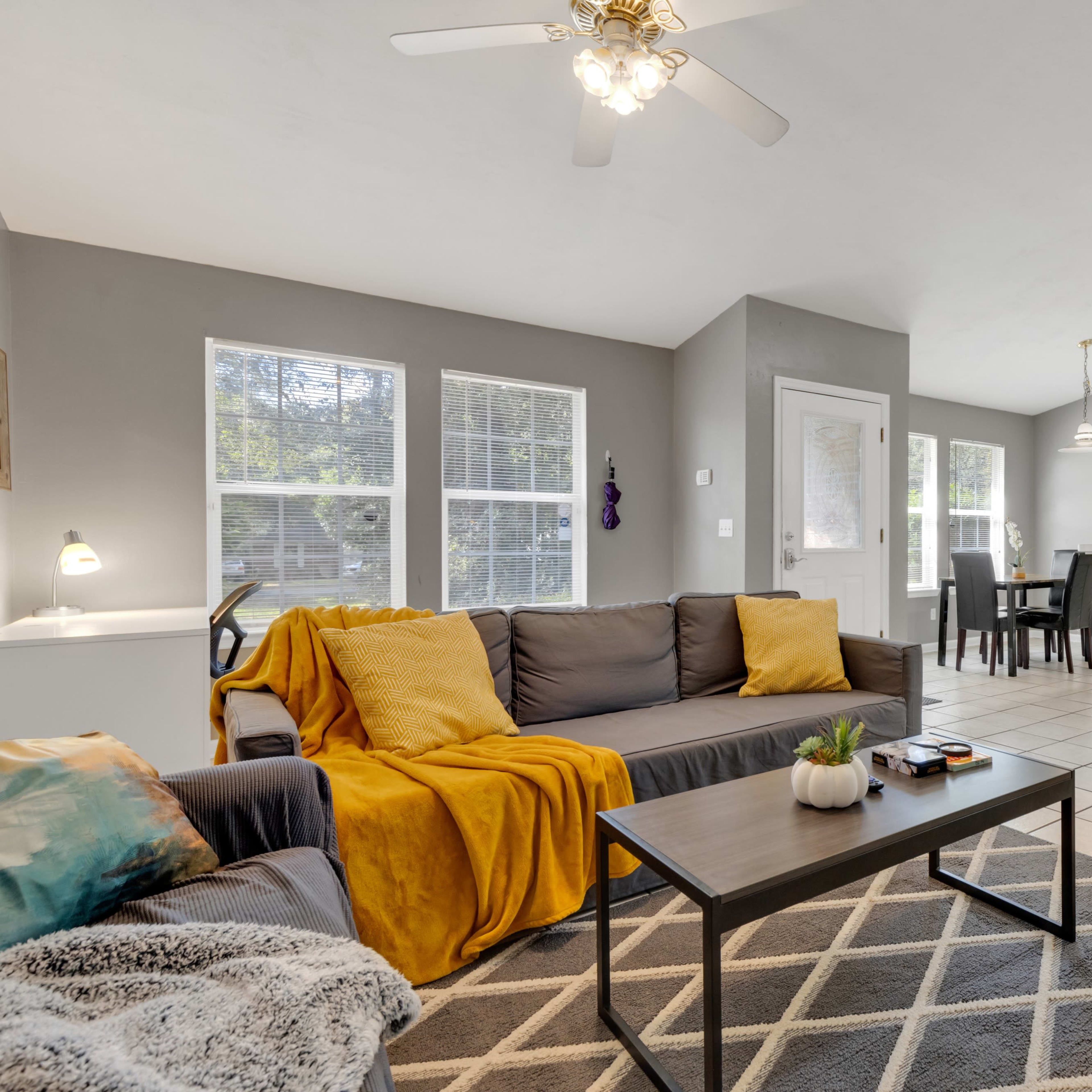 A living room with a grey couch and yellow throw blanket, a coffee table, and an open kitchen area in a well-lit, modern space.