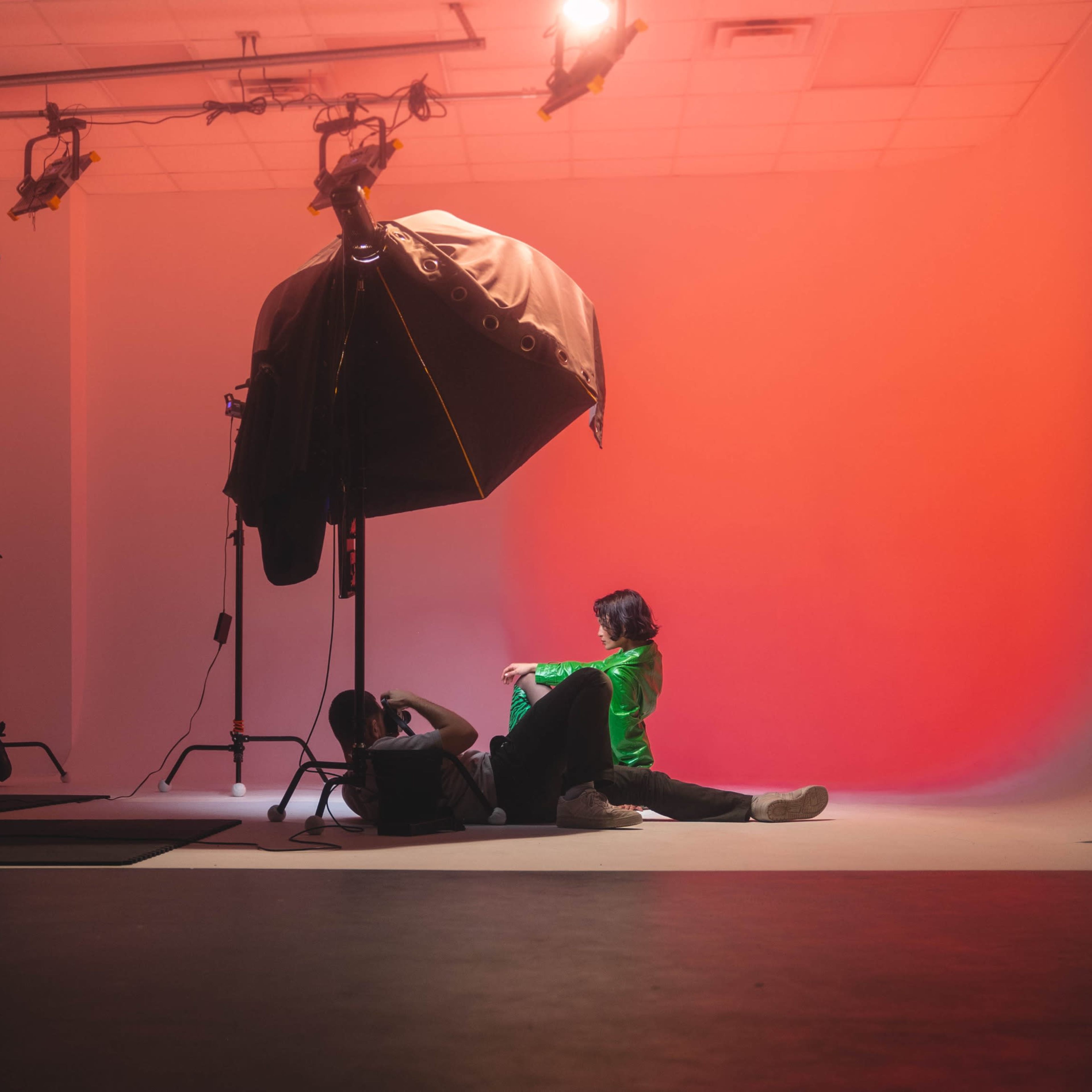 A person wearing a green jacket is seated on the floor of a photography studio with a large softbox lighting setup and a gradient backdrop.