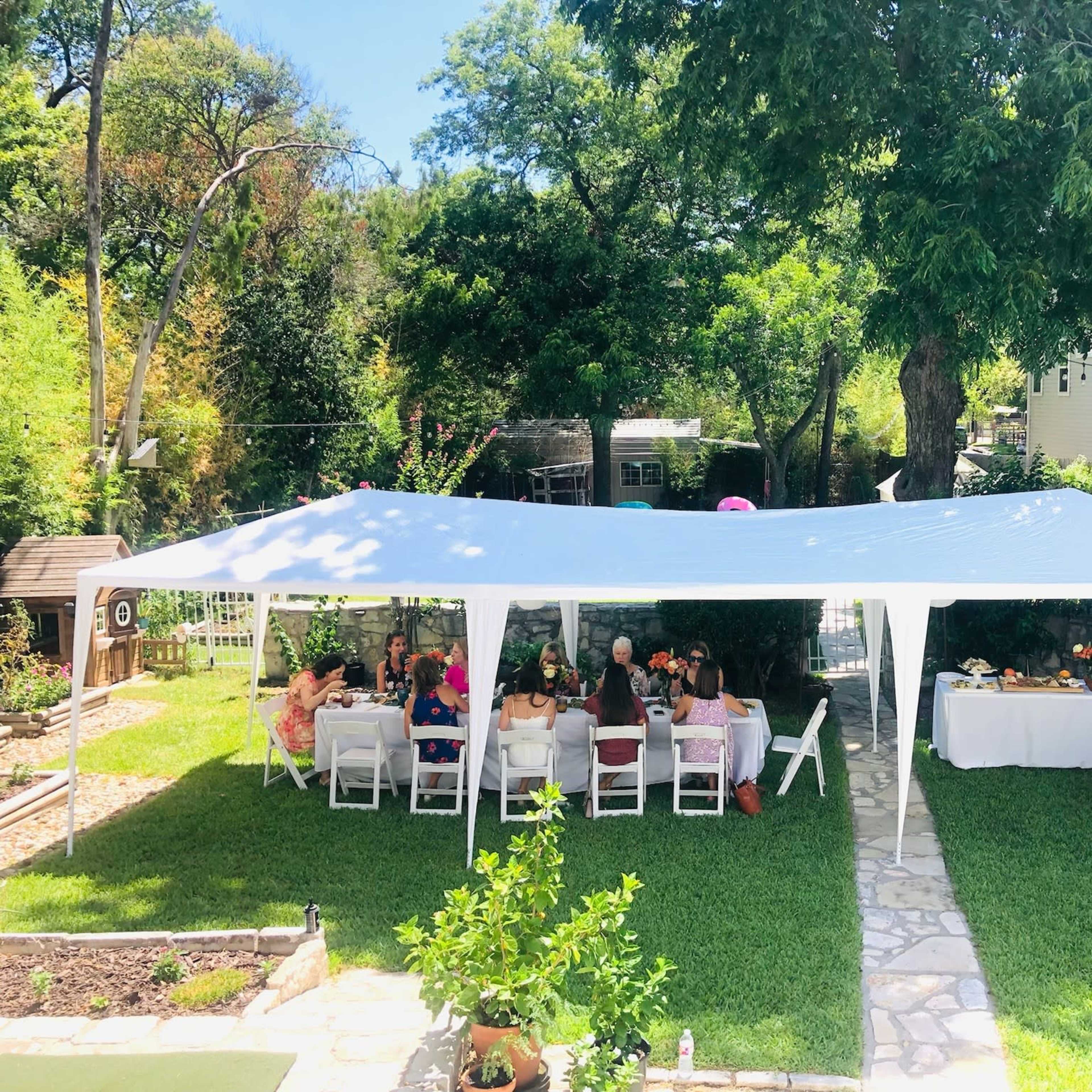A large white tent is set up in a green backyard, where guests are seated at tables adorned with floral centerpieces.