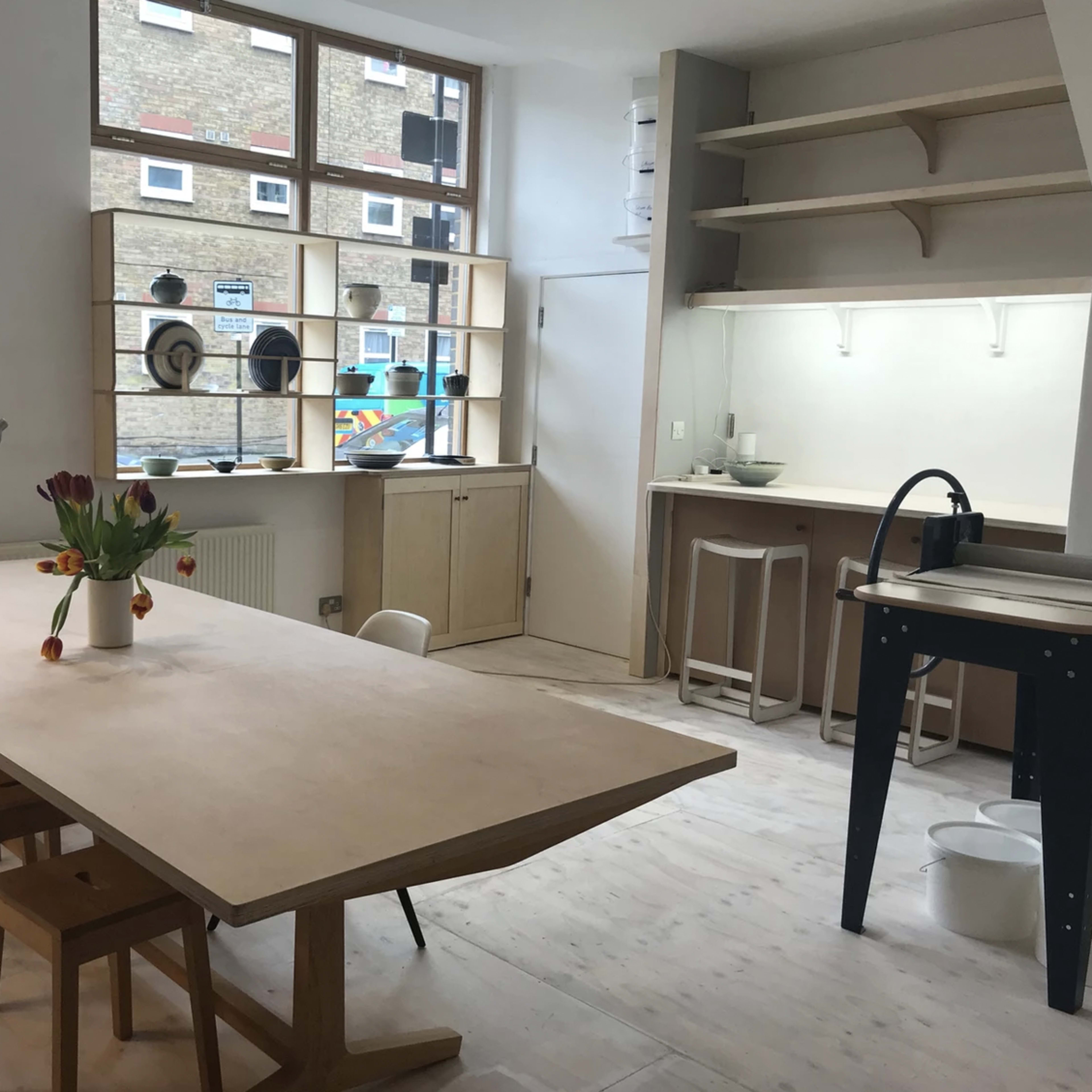 A minimalist modern interior with a long wooden dining table, chairs, and shelving displaying decorative items.