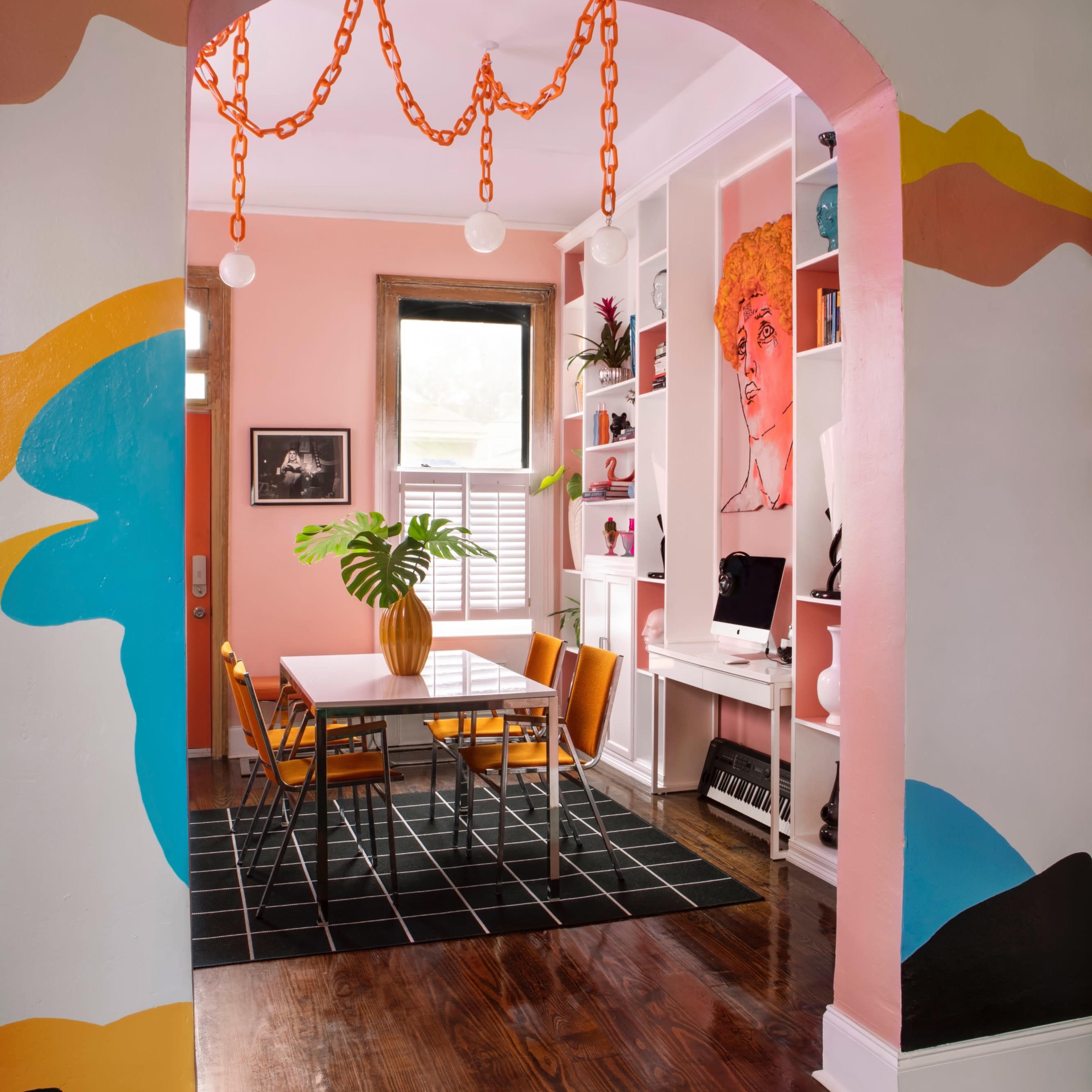 A brightly decorated room features a dining area with a white table and orange chairs, framed by an archway with colorful wall art and a plant centerpiece.