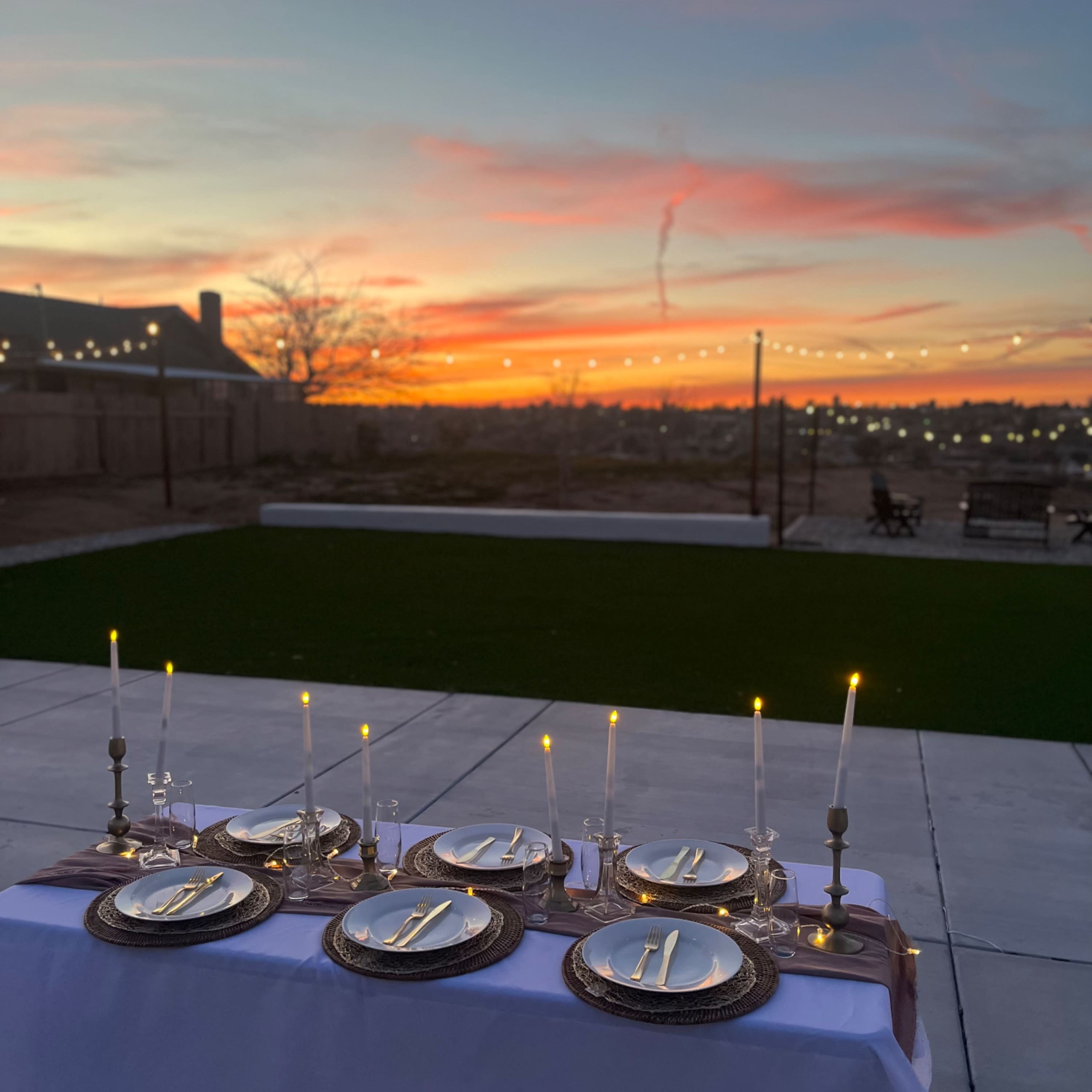 A neatly set outdoor dining table with plates, cutlery, and candles is positioned against a vibrant sunset sky.