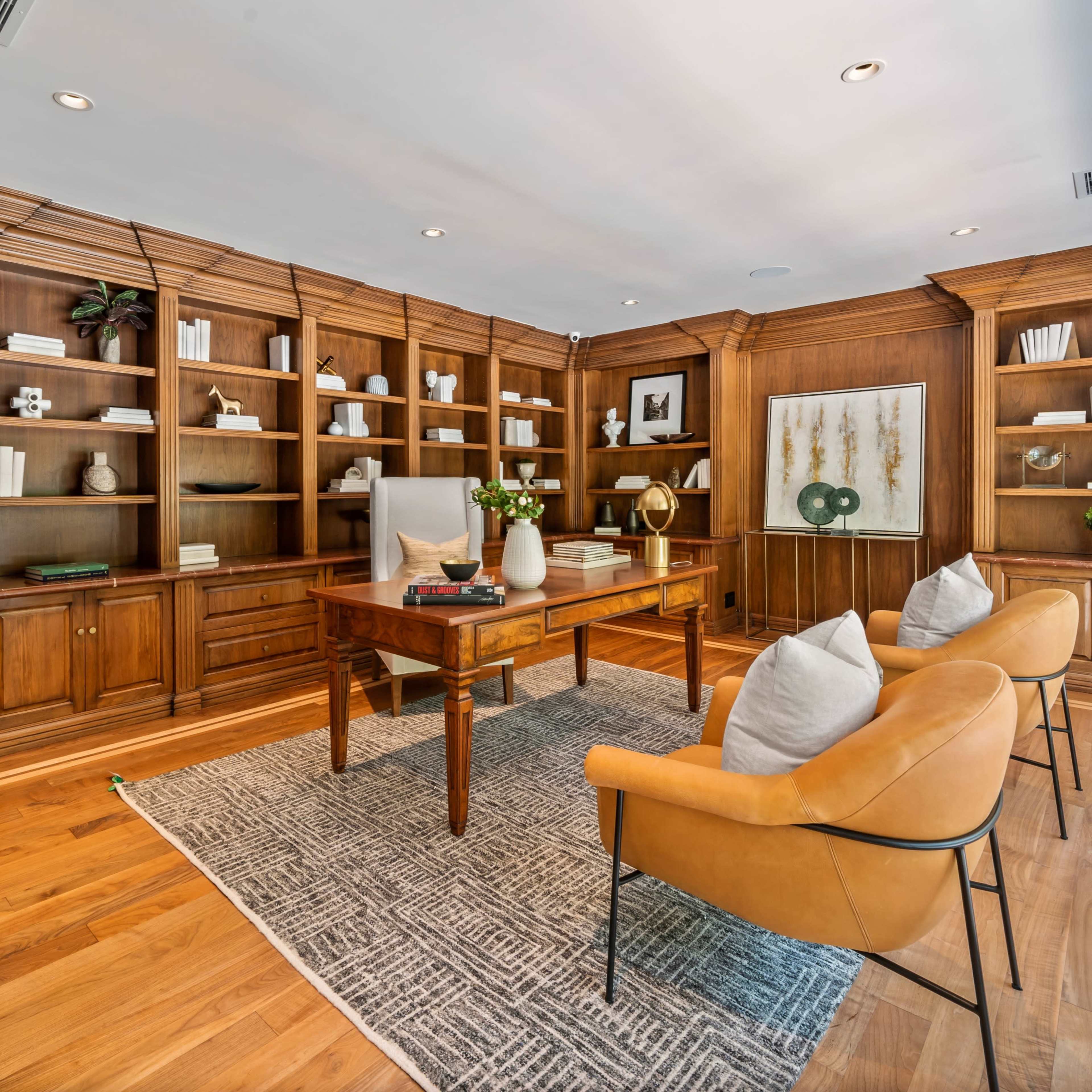A wooden office space features built-in bookshelves, a central desk, and two accent chairs on a patterned rug.