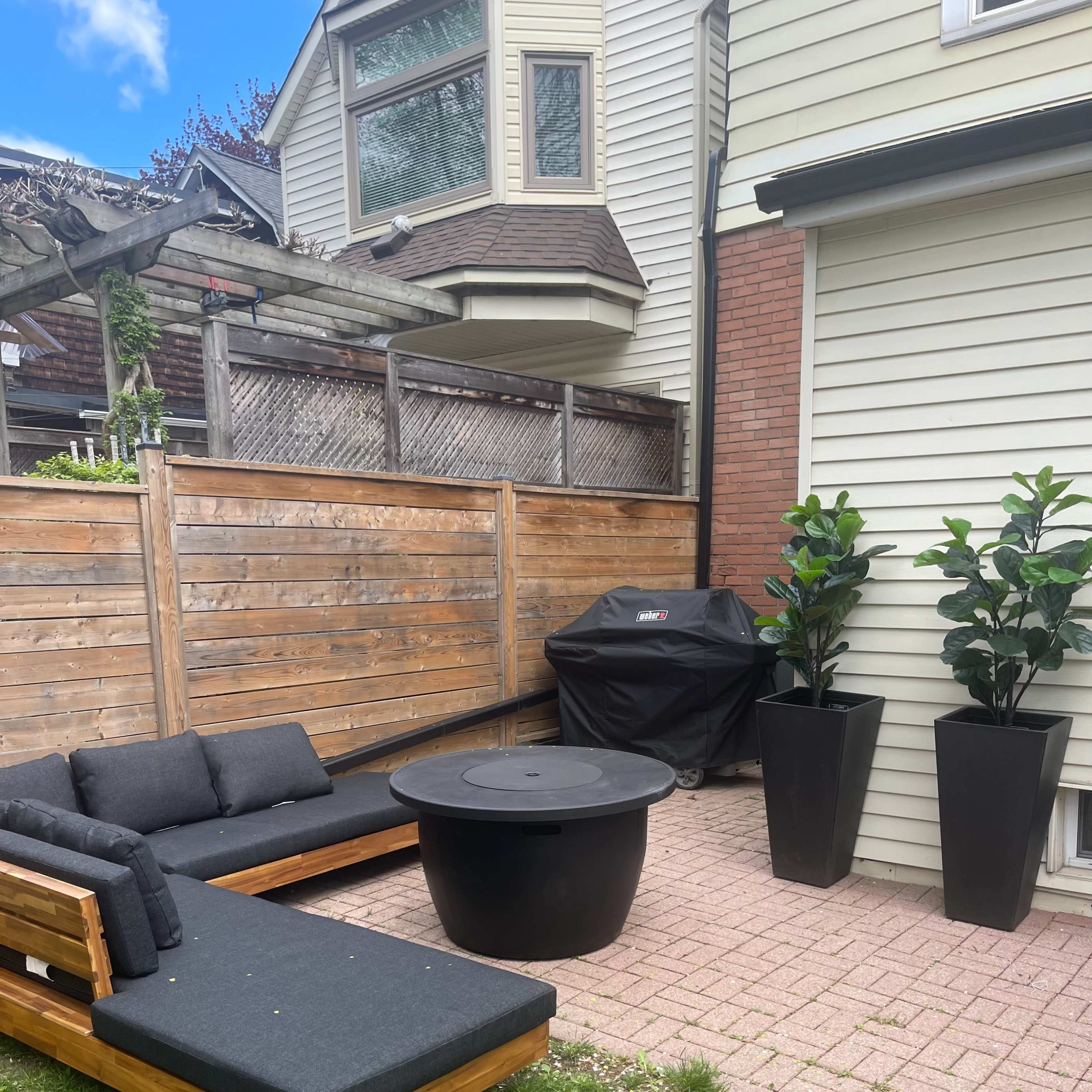 A patio area with a wooden seating arrangement, a round black table, a grill, and tall planters with plants, enclosed by a wooden fence.